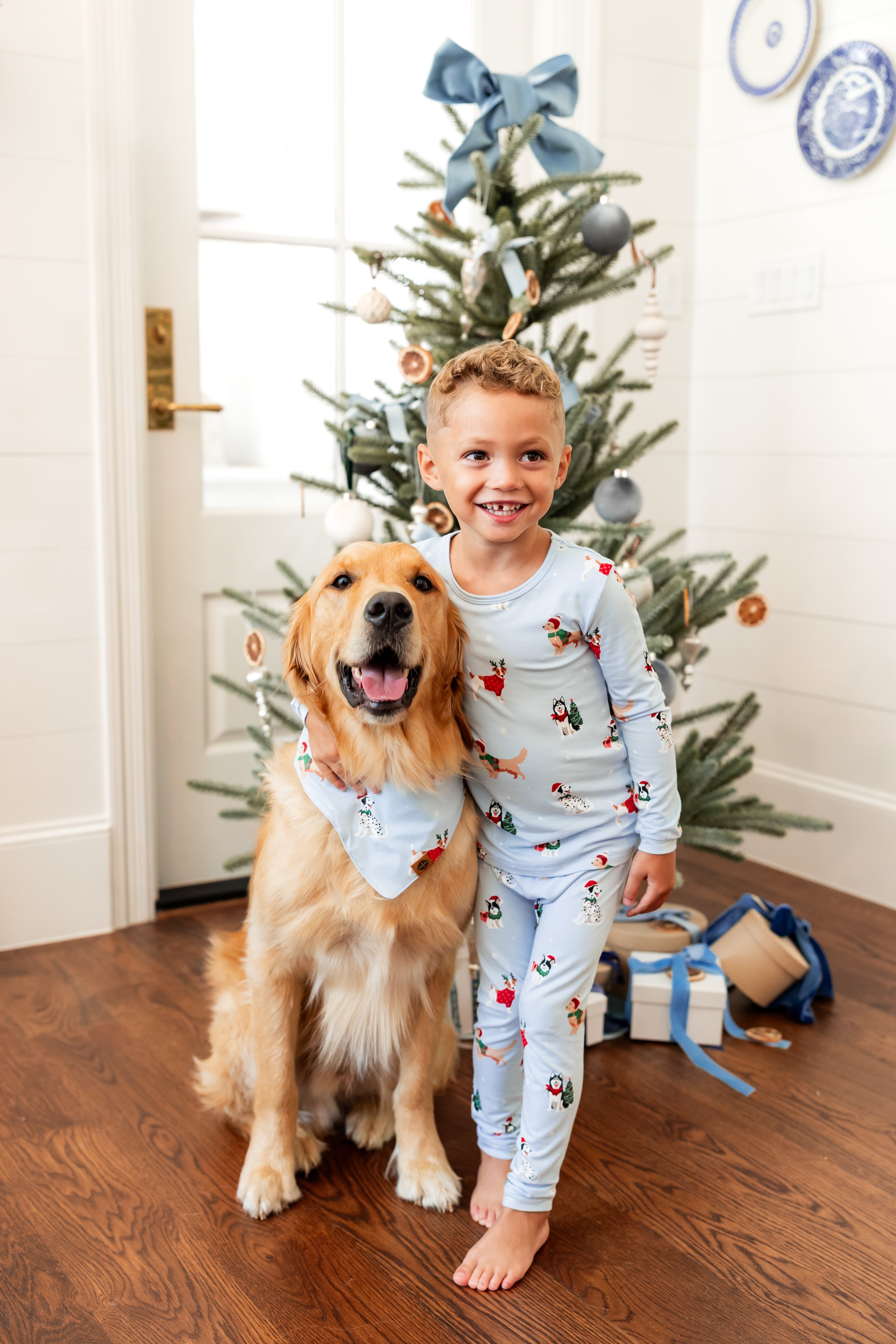 Boy wearing Long Sleeve Pajamas in Holiday Pups print while standing with a dog wearing similar print dog bandana with Christmas tree and presents in the background