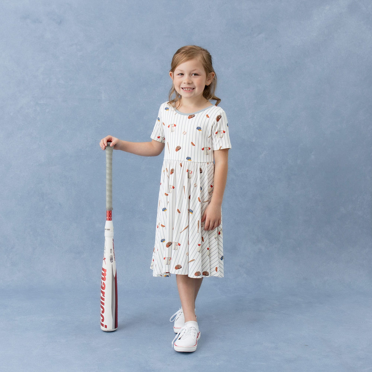 Young girl wearing the Twirl Dress in Home Run standing beside a baseball bat
