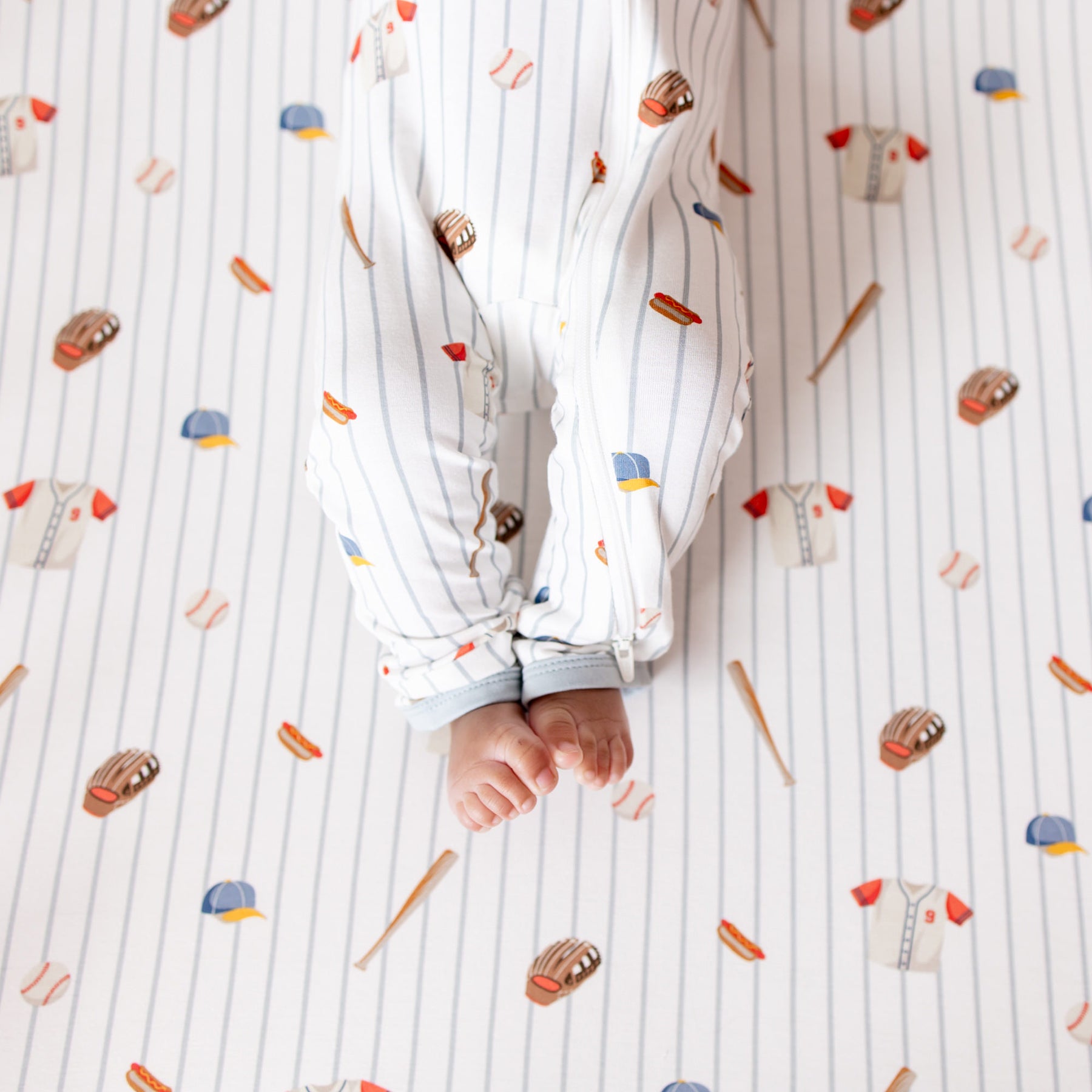 The zipper closure on the leg of the Zippered Romper in Home Run shown on an infant model laying on a Crib Sheet in Home Run