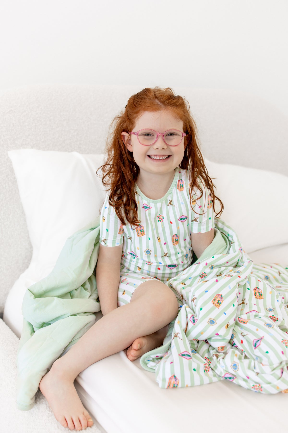 Child sitting on a bed wrapped in a green and white striped youth blanket with colorful Harry Potter patterns wearing a similar print short sleeve pajama set
