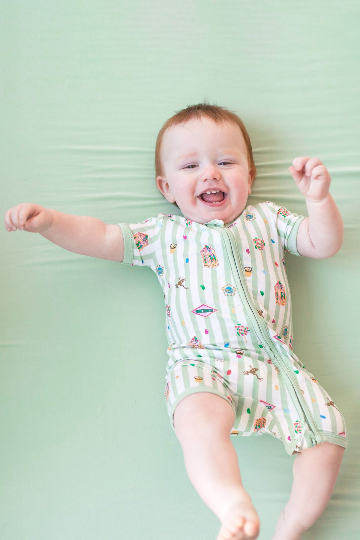 Baby wearing a green and white striped zippered shortall romper with colorful Harry Potter patterns lying on a green background