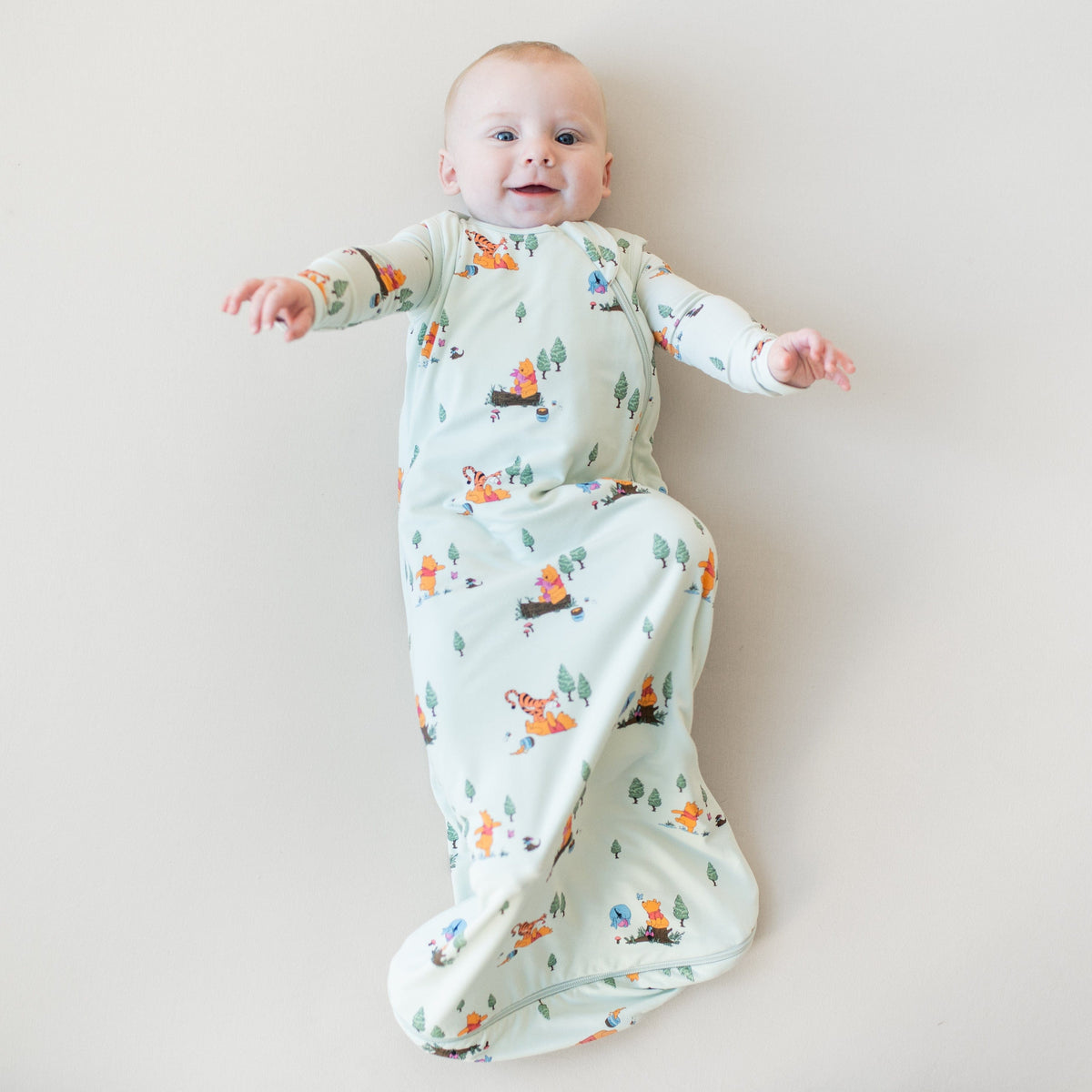 Infant laying on a light neutral surface wearing the Sleep Bag in Winnie the Pooh Hundred Acre Wood 0.5 with a matching romper