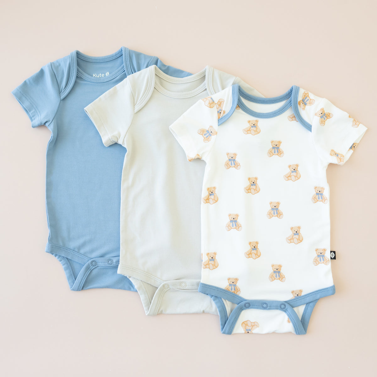 Three baby bodysuits in blue, off-white, and patterned with teddy bears on a beige background.