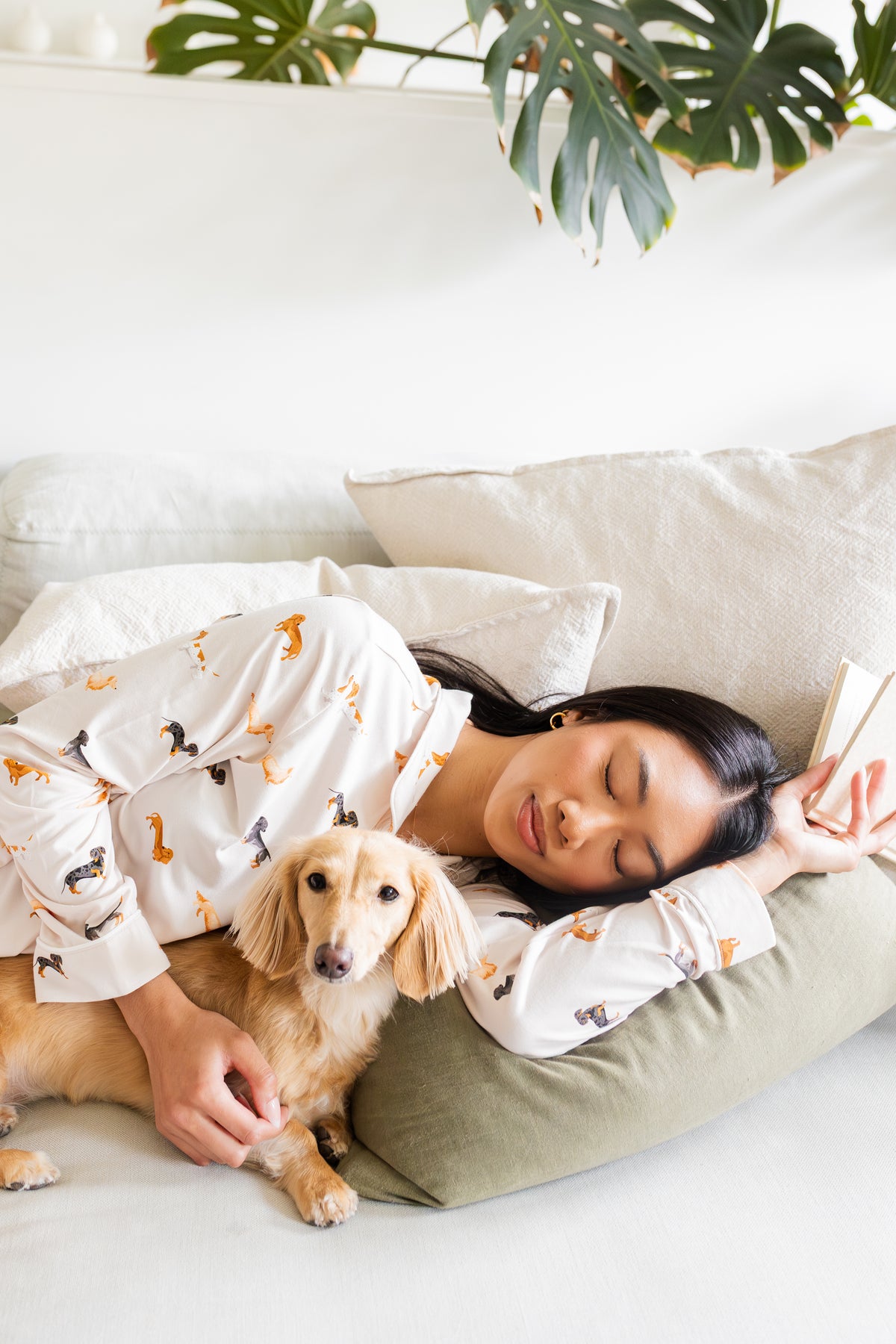Woman in dachshund dog patterned pajamas lying on a couch with a small dog, surrounded by plants