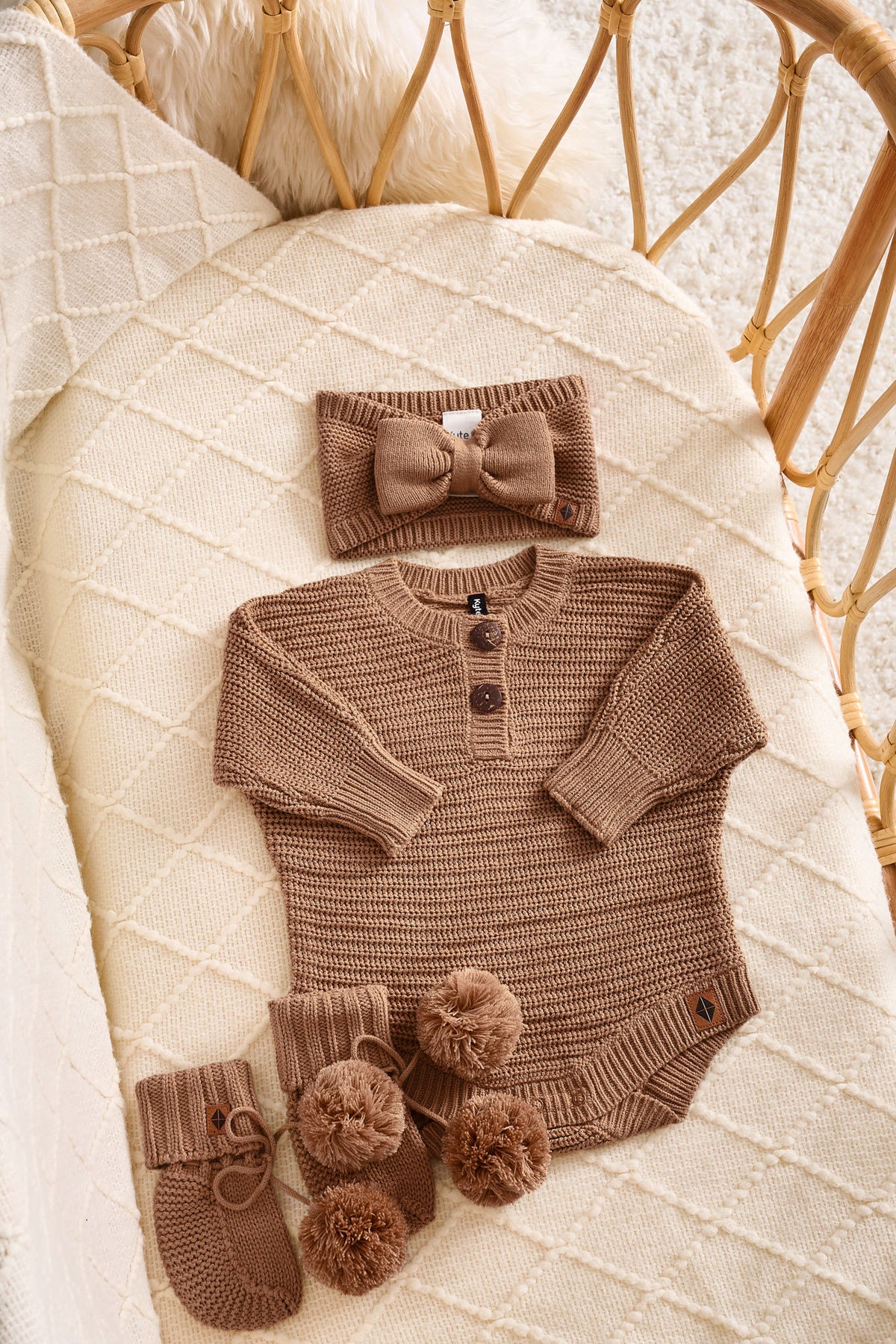 A baby knit outfit set in a warm taupe-brown color, including a long-sleeve henley bodysuit with three buttons, a matching bow headband, and two booties with pom-poms, arranged on a cream quilted blanket inside a rattan bassinet.