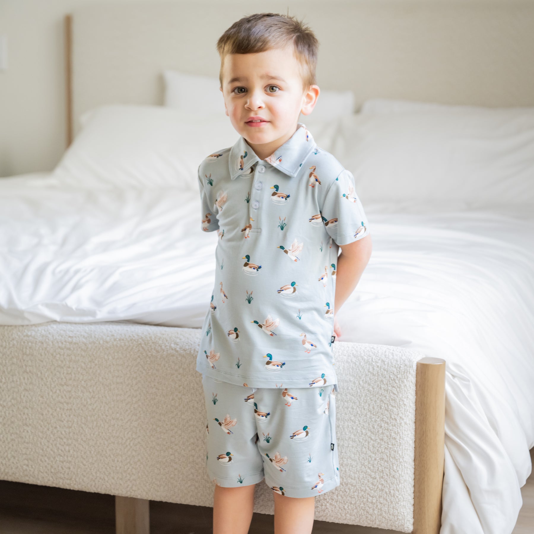 Young boy standing at the end of a bed wearing the Toddler Short Sleeve Polo in Mallard Duck with matching drawstring shorts