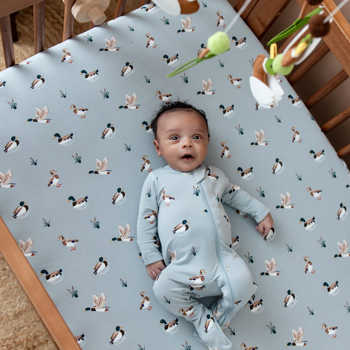Infant wearing the Zippered Footie in Mallard Duck laying in a crib on a mallard duck crib sheet