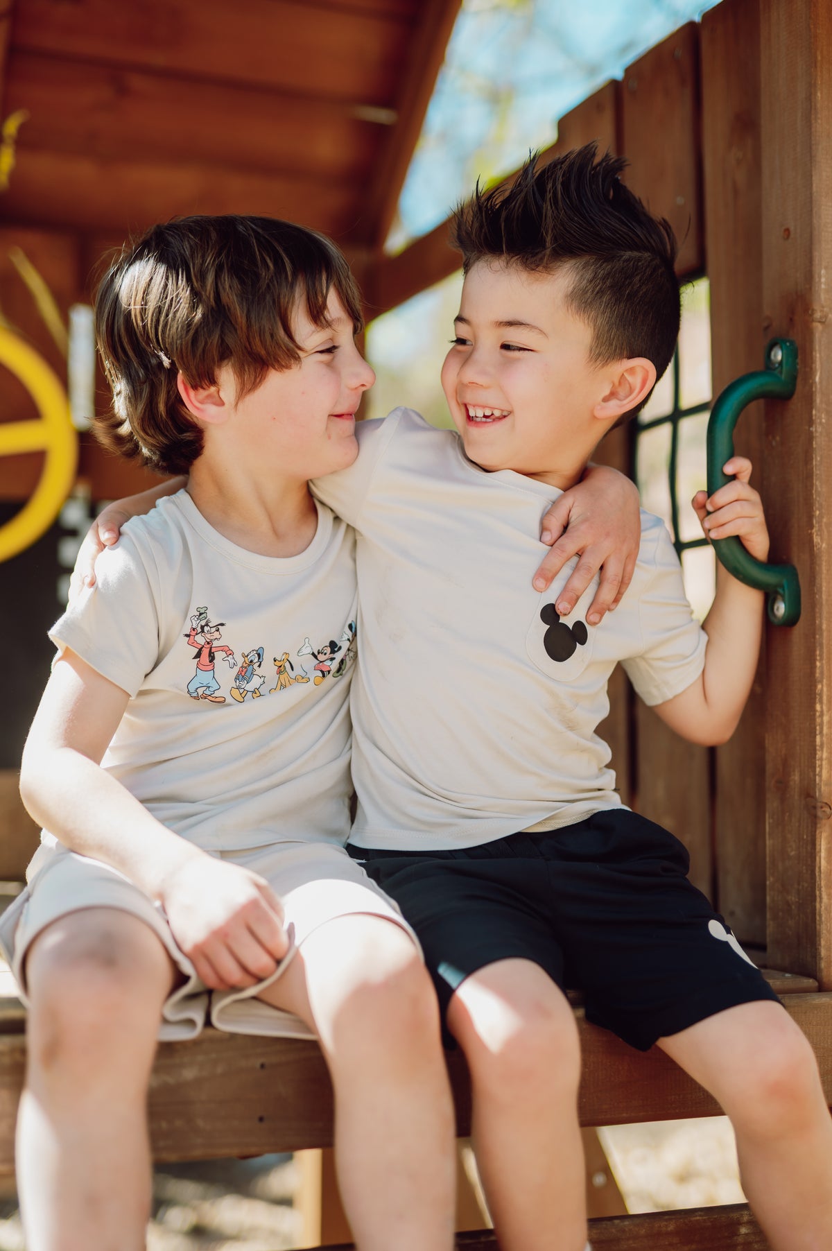 Two children sitting on a wooden structure, smiling and hugging.