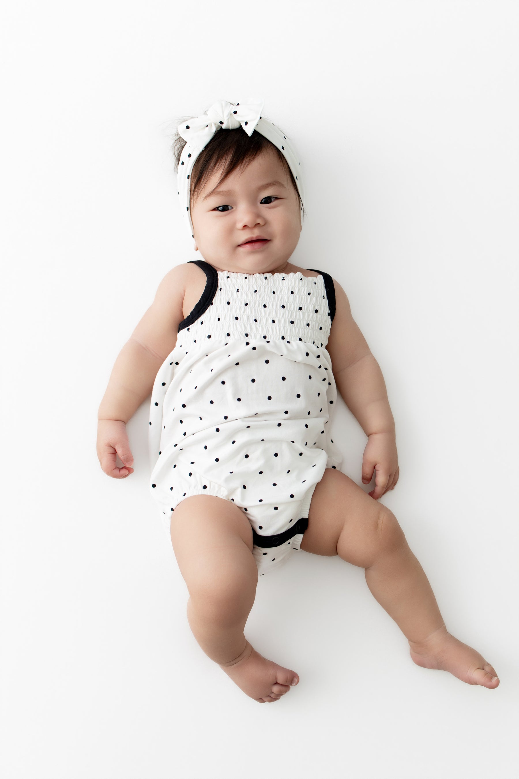 Baby wearing Smocked Bubble Romper and bow headband with Midnight Polka Dot print on a white background