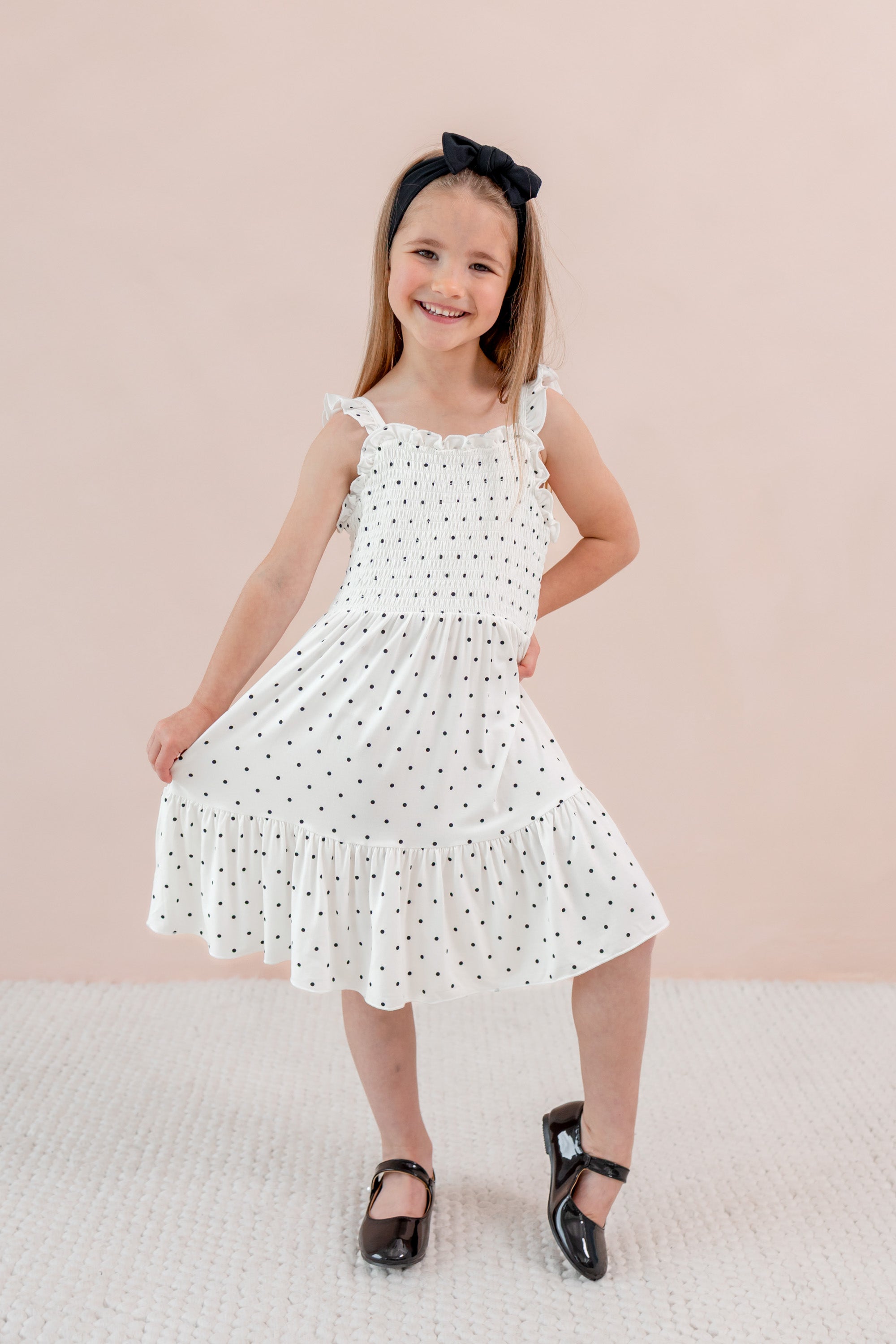 Child wearing Smocked Dress in Midnight Polka Dot print with Black bow headband and shoes on a beige background