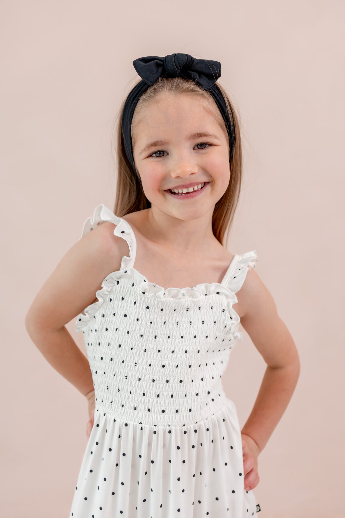 Child smiling in Smocked Dress in Midnight Polka Dot print with Black bow headband on a beige background
