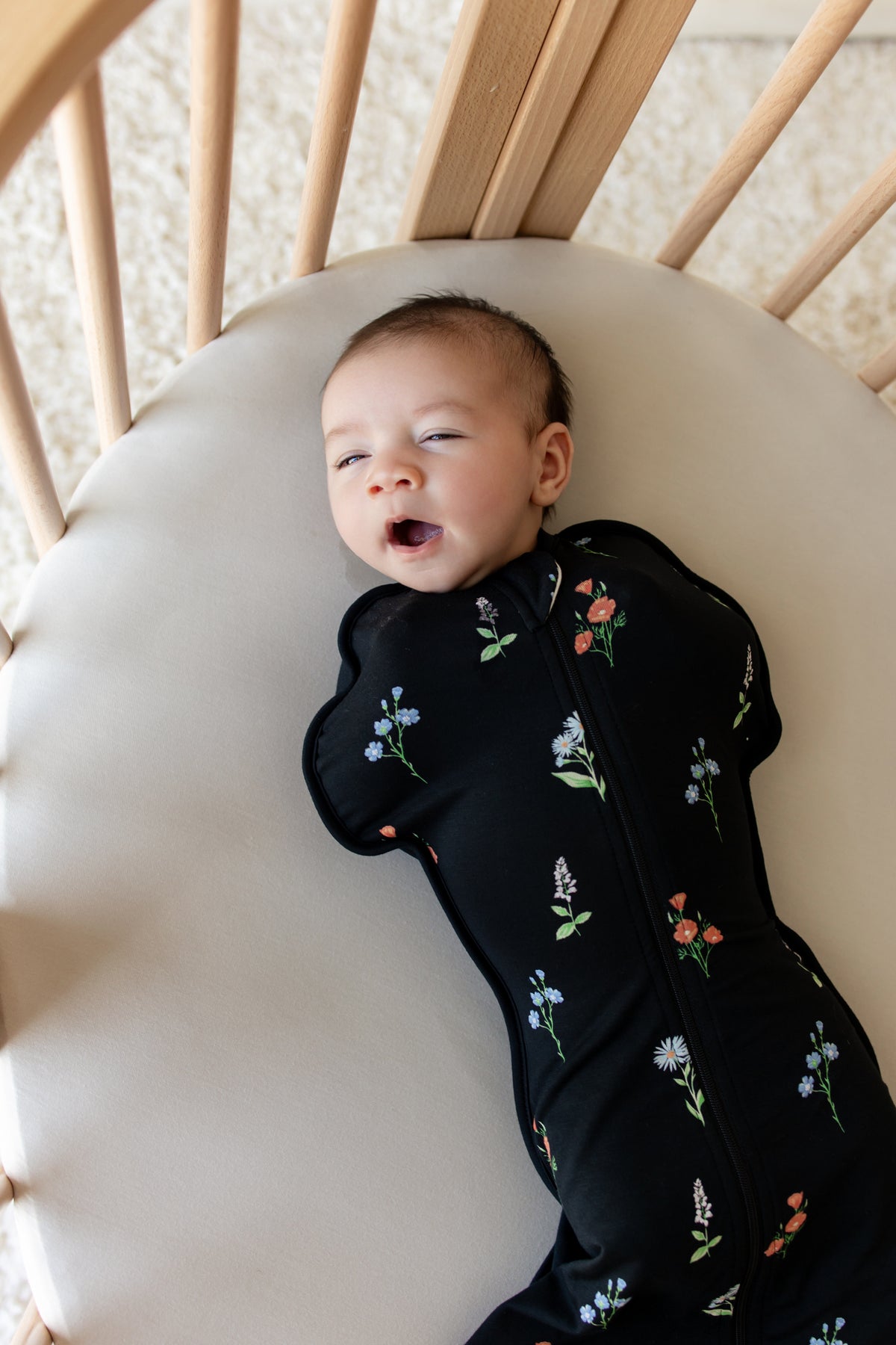 Baby yawning in a crib wearing black Swaddle Bag with Wildflower print