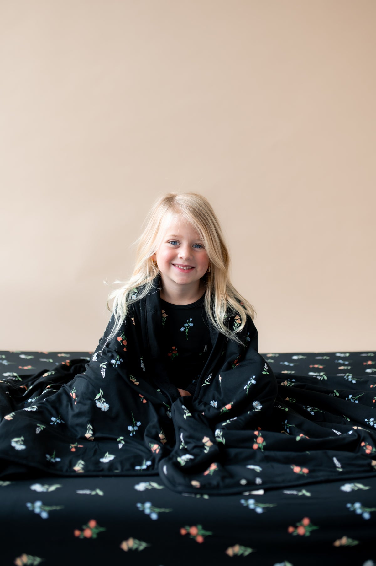A girl wrapped in Toddler Blanket in Midnight Wildflower 1.0 print sitting on a similar print twin sheet and wearing similar print pajamas on a beige background