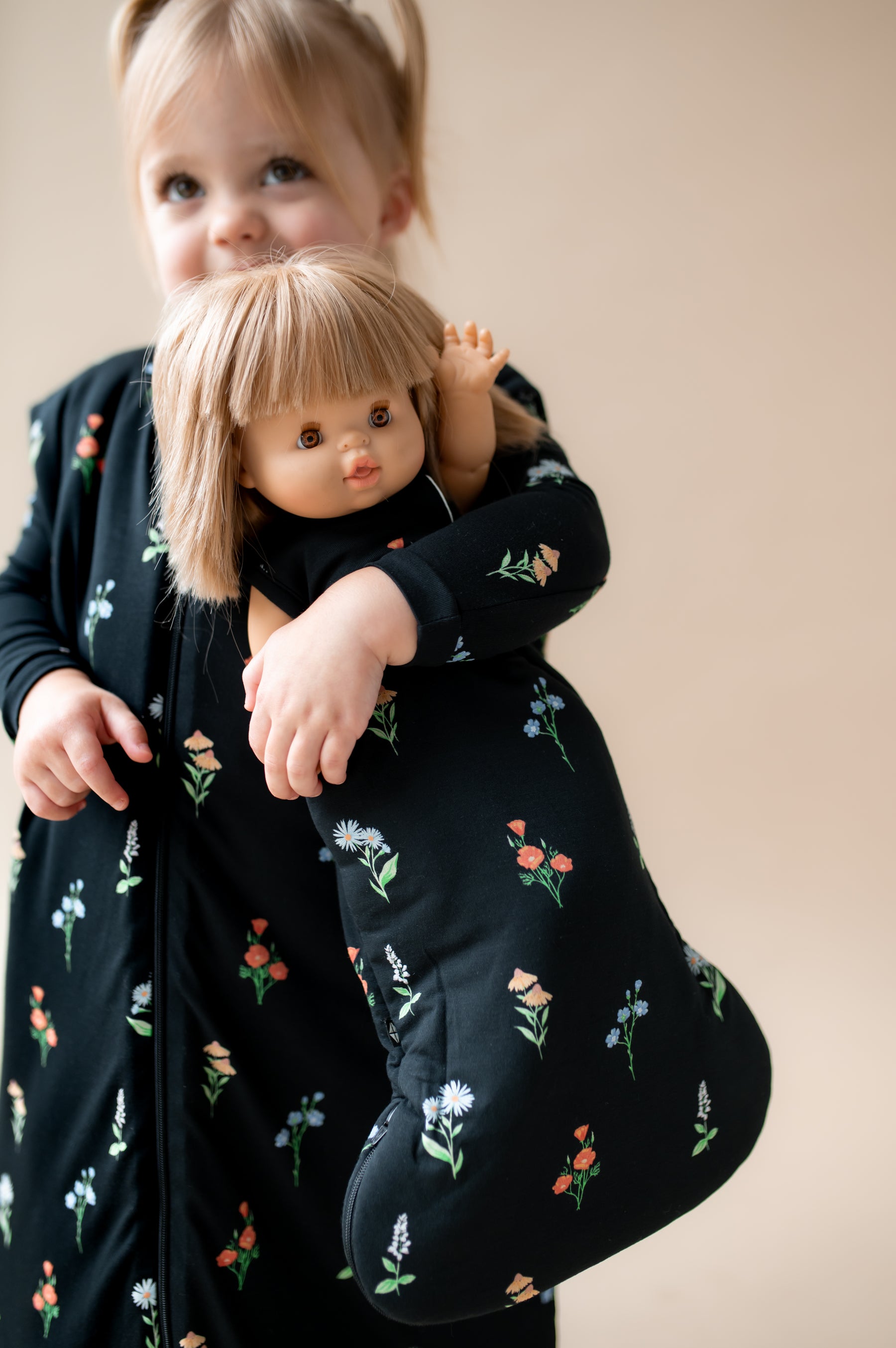 Girl holding a baby doll sleep bag in midnight wildflower while also wearing Sleep Bag Walker in Midnight Wildflower 2.5 print on a beige background