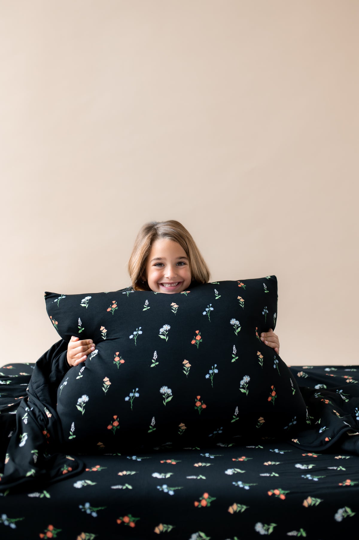 Girl holding Standard Pillowcase in Midnight Wildflower print sitting on a similar print sheets and youth blanket on a beige background
