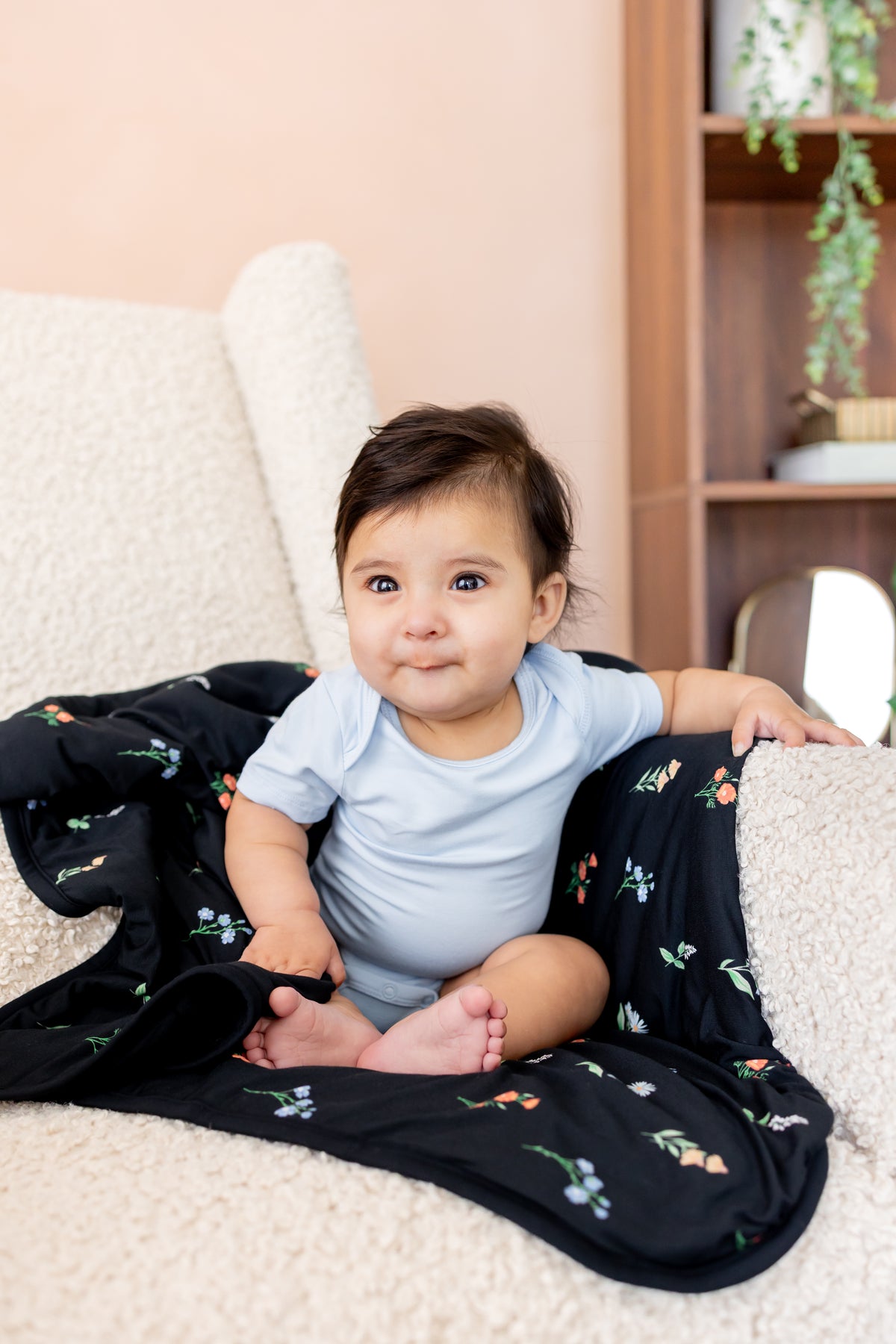 Baby sitting on a white sofa with Baby Blanket in Midnight Wildflower 1.0 print wearing a blue bodysuit