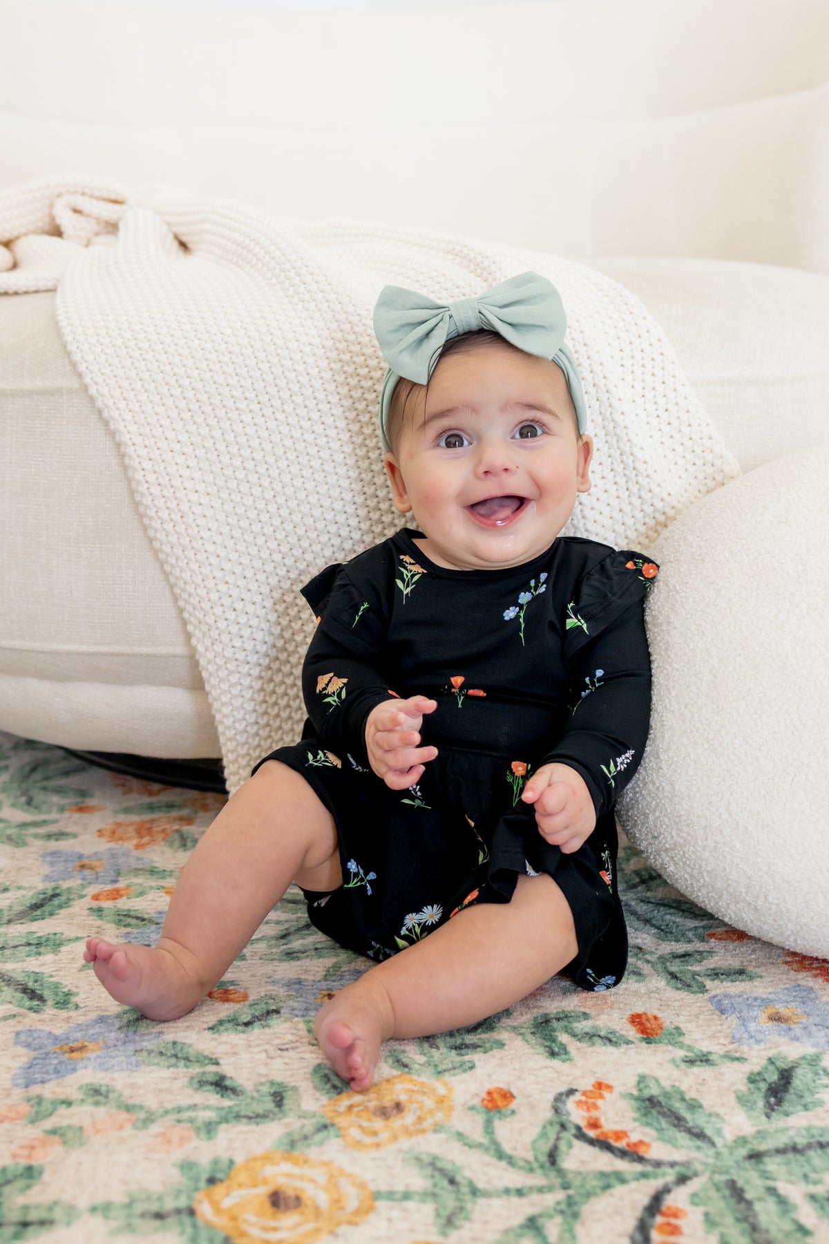 Baby sitting against a white couch smiling in Long Sleeve Twirl Bodysuit Dress in Midnight Wildflower print and a blue head bow
