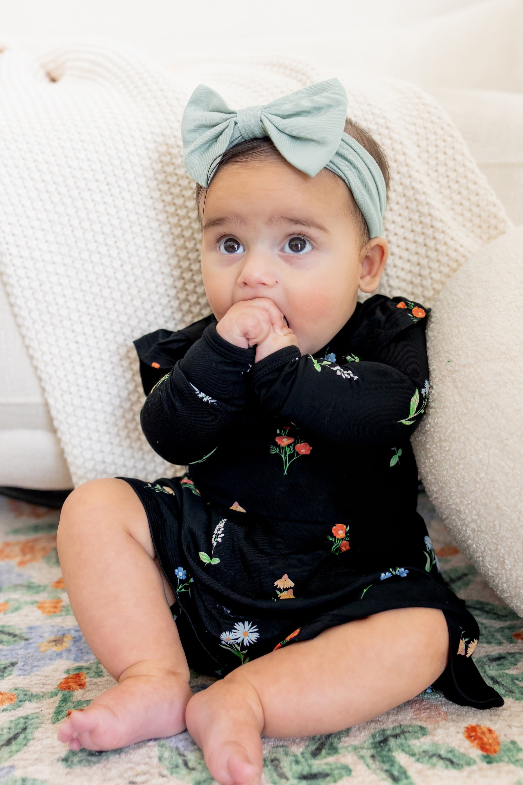 Baby sitting against a white couch wearing Long Sleeve Twirl Bodysuit Dress in Midnight Wildflower print and a blue head bow