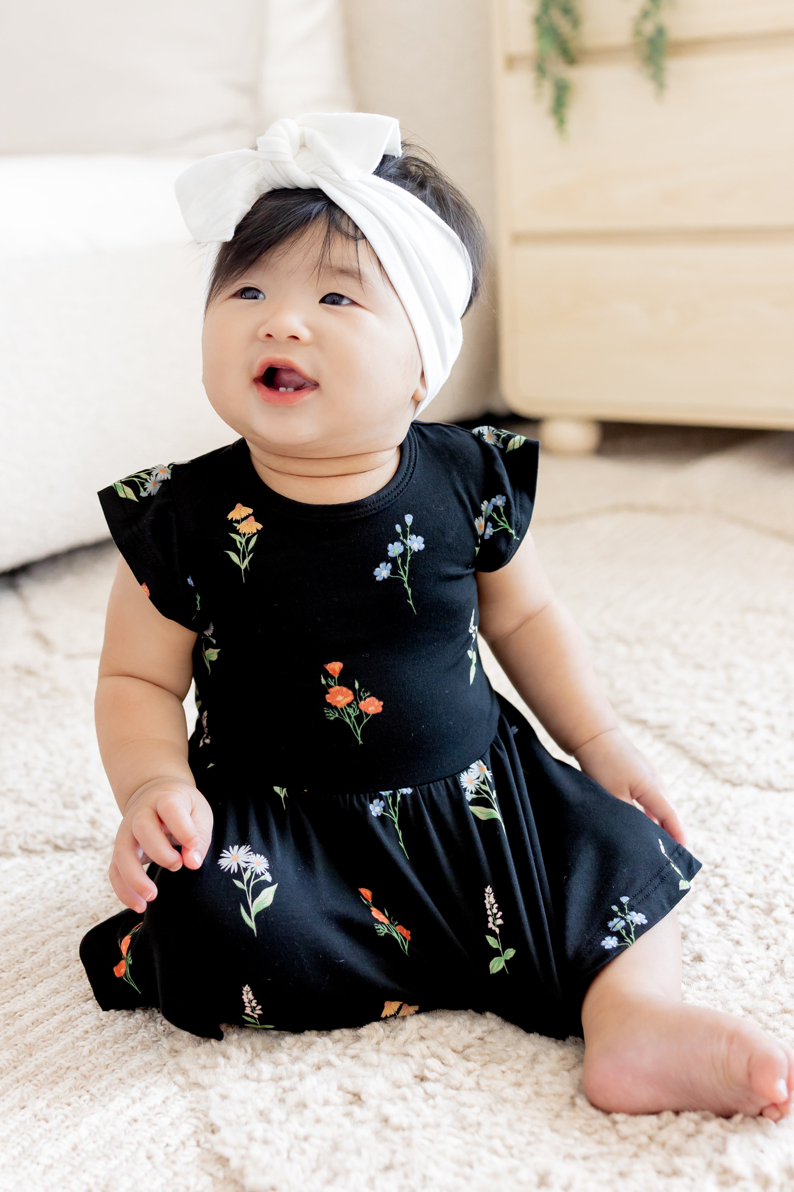 Baby sitting in Twirl Bodysuit Dress in Midnight Wildflower print wearing a white head bow on a white carpet