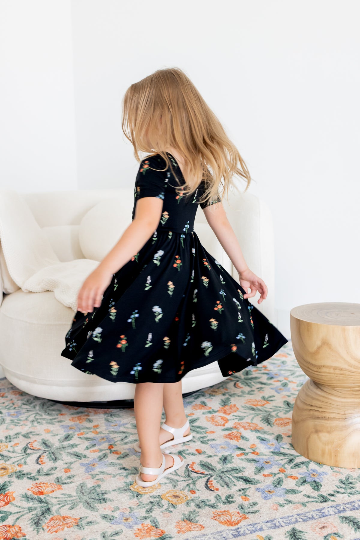 Girl twirling in Twirl Dress in Midnight Wildflower print and white sandals standing in a living room