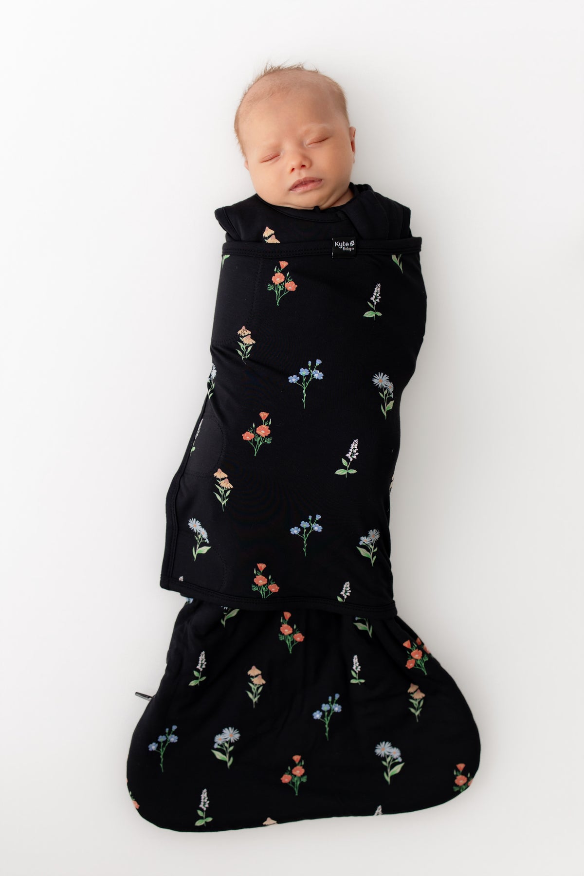 Baby sleeping in Sleep Bag Swaddler in Midnight Wildflower 1.0 print on a white background