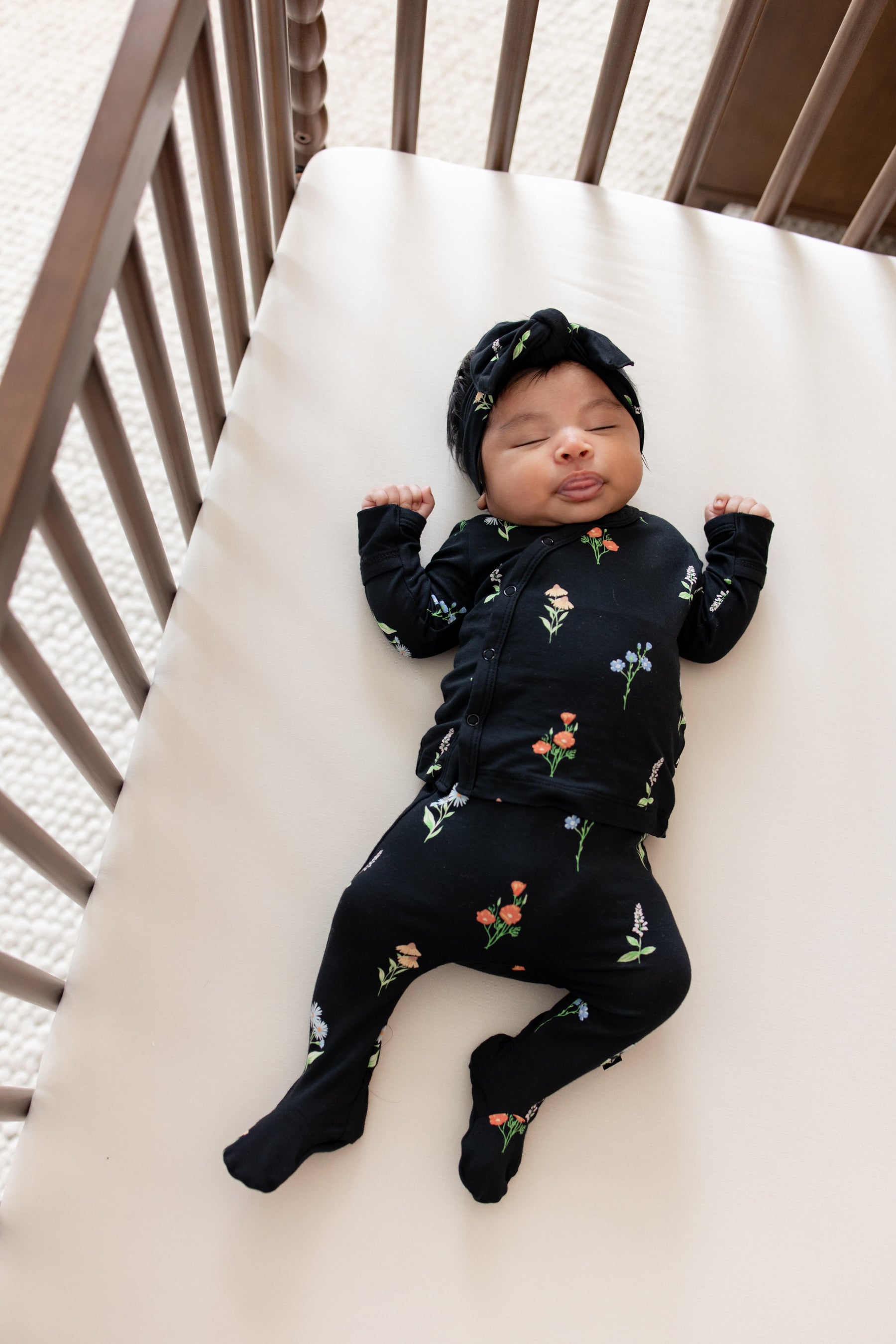 Baby sleeping in a crib wearing Take Me Home Set with Bow in Midnight Wildflower print lying on beige crib sheet.