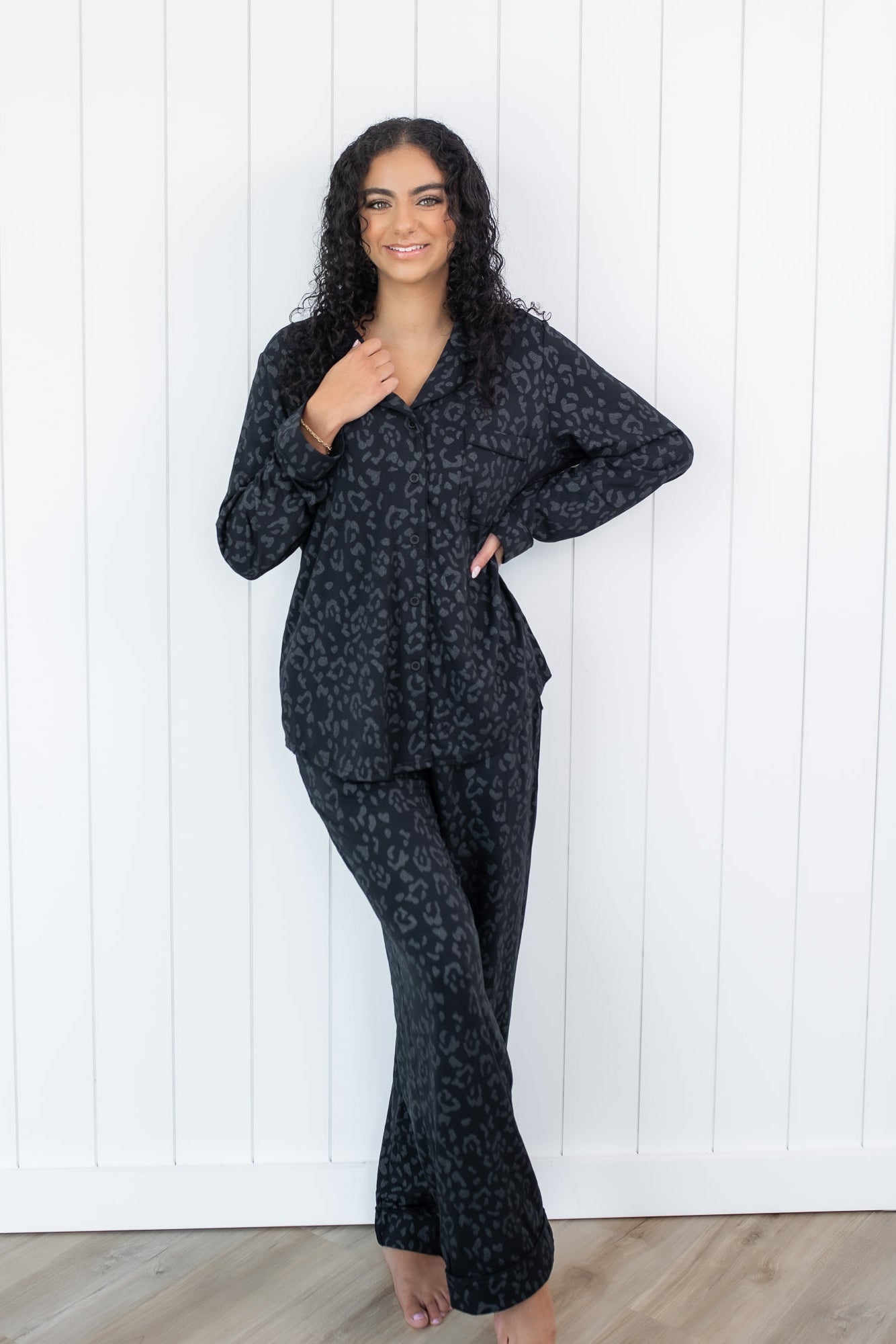 Female standing in front of a white paneled wall wearing the Long-Sleeved Women's Pajama Set in Midnight Leopard