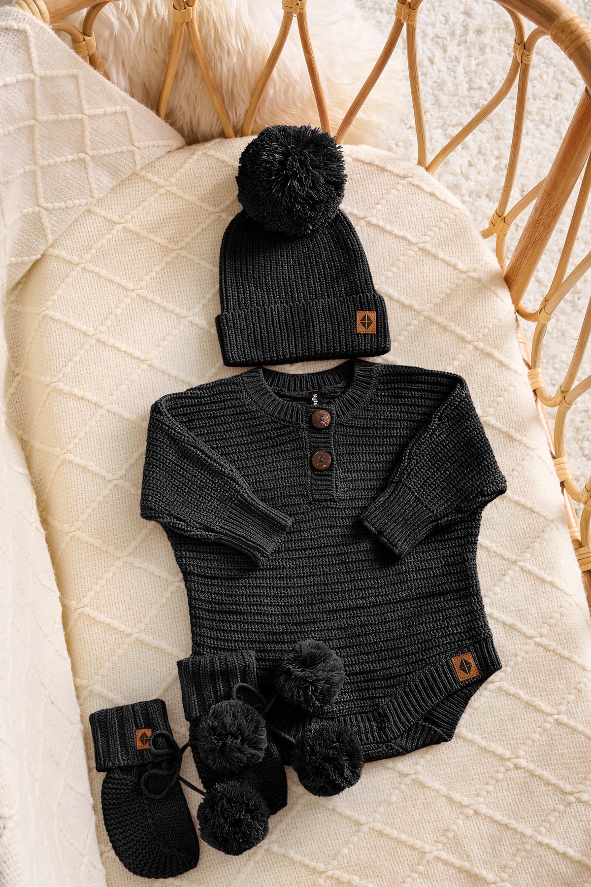 A baby knit outfit set laid flat on a cream quilted blanket, including a black long-sleeve henley bodysuit with brown buttons, a matching black beanie with a pom-pom and small brown logo tab, and black booties with pom-poms. A woven rattan chair is visible in the background.