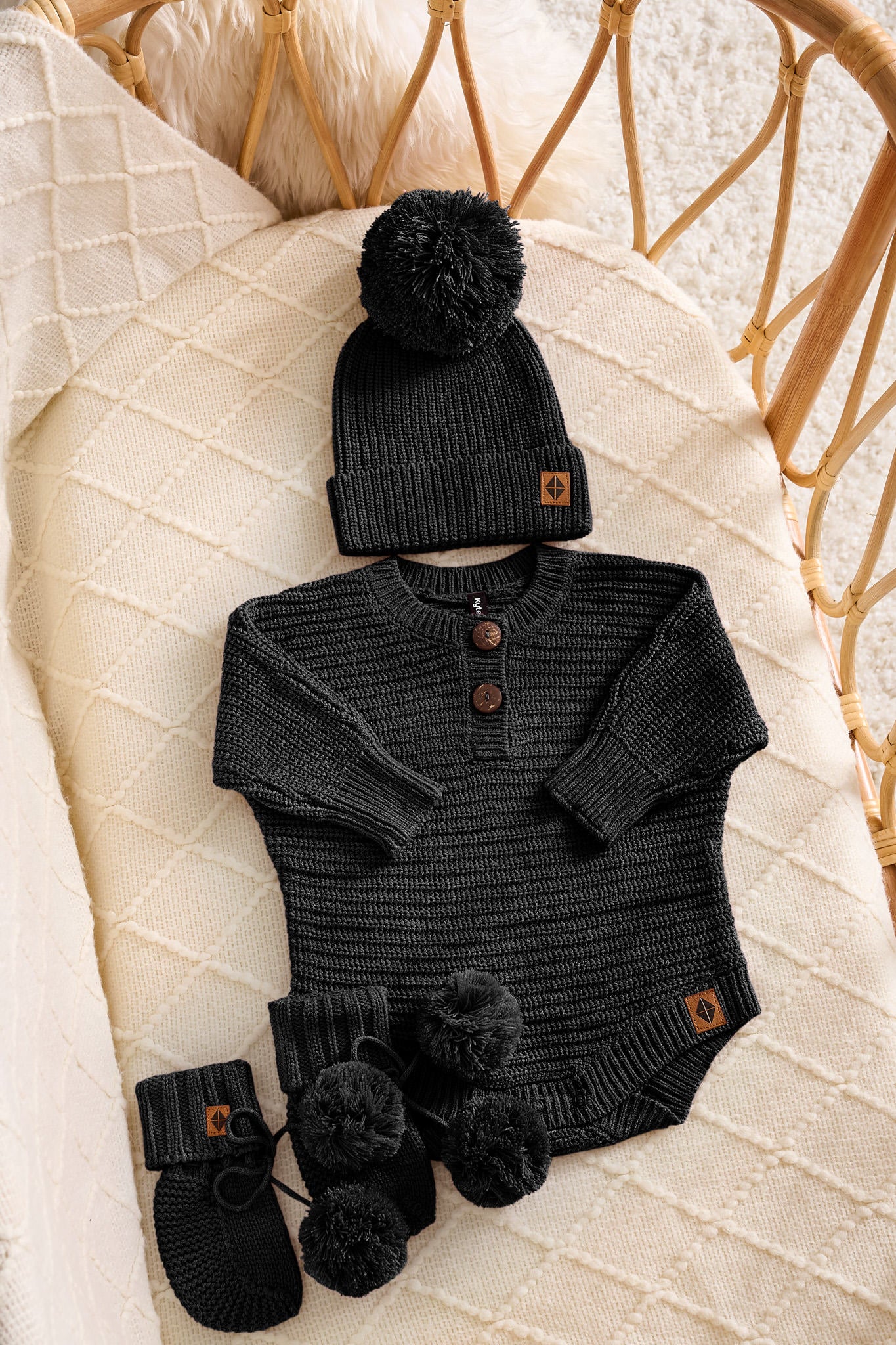 A baby knit outfit set laid flat on a cream quilted blanket, including a black long-sleeve henley bodysuit with brown buttons, a matching black beanie with a pom-pom and small brown logo tab, and black booties with pom-poms. A woven rattan chair is visible in the background.