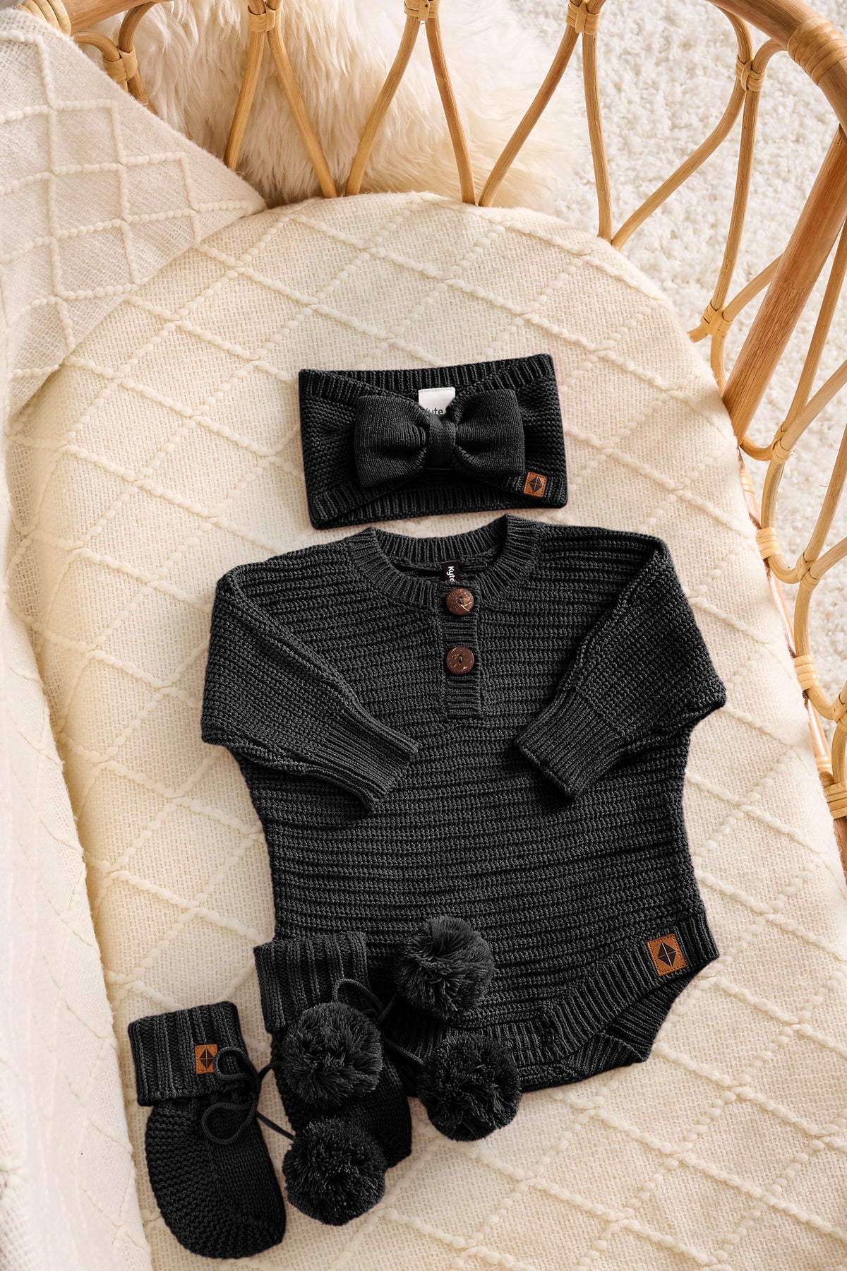 A baby clothing set laid flat on a cream quilted blanket, including a black ribbed long-sleeve bodysuit with three brown buttons, a matching black bow headband, black booties with pom-poms, and a small brown brand tab. A woven rattan chair is visible in the background.
