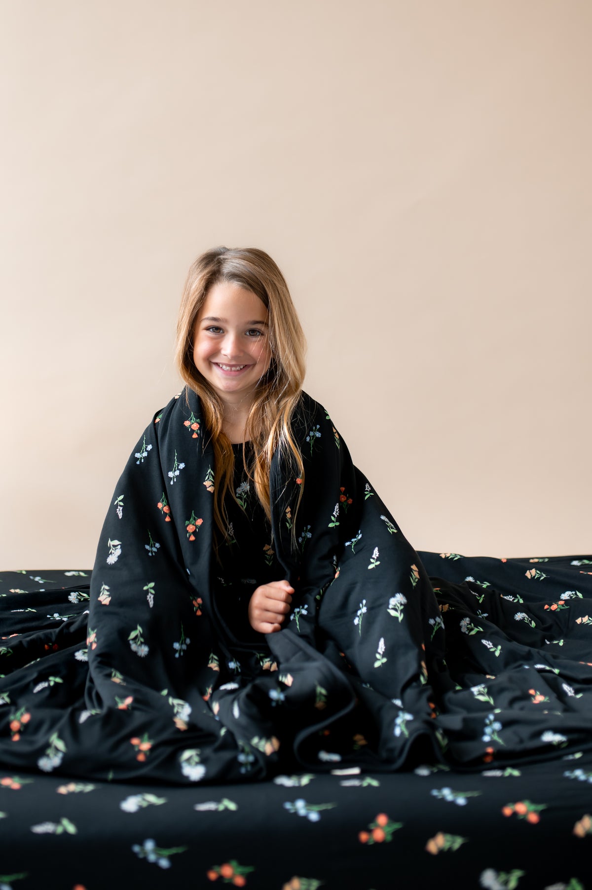 Girl wrapped in Youth Blanket in Midnight Wildflower 2.5 print sitting on a similar print sheet on a beige background