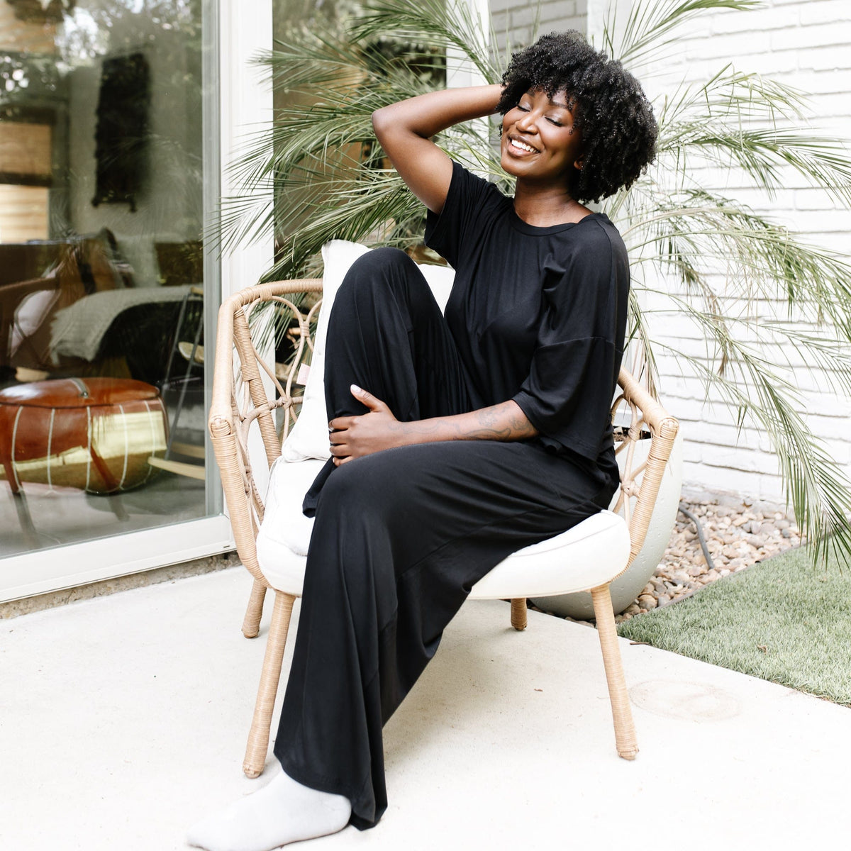 Smiling model sitting with her eyes closed in a wicker chair with white cushions wearing the Women's Short Sleeve Tee in Midnight with matching wide leg pants