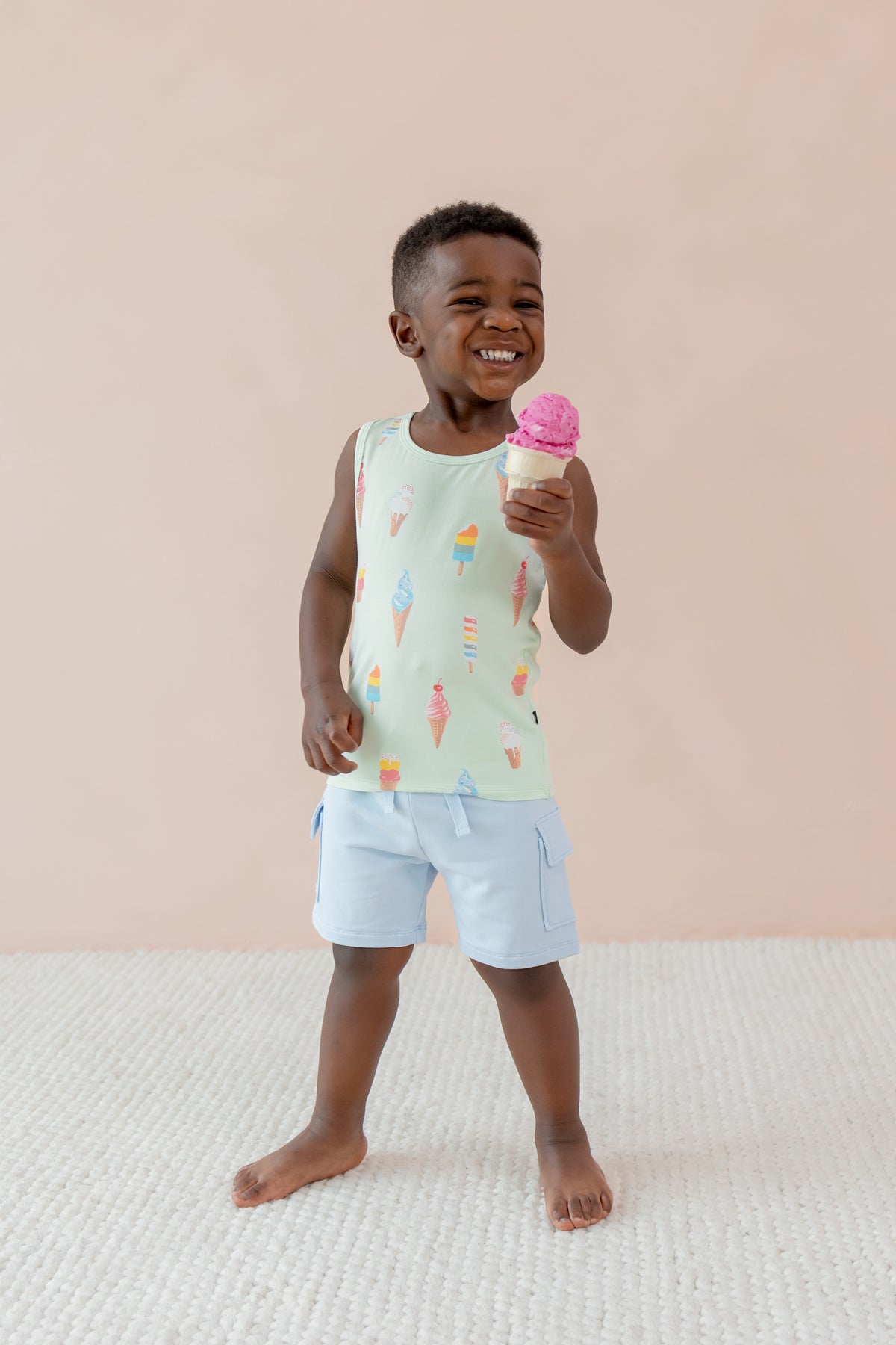 Child smiling and holding an ice cream cone wearing a Toddler Tank Top in Mint Ice Cream print over blue shorts on a beige background