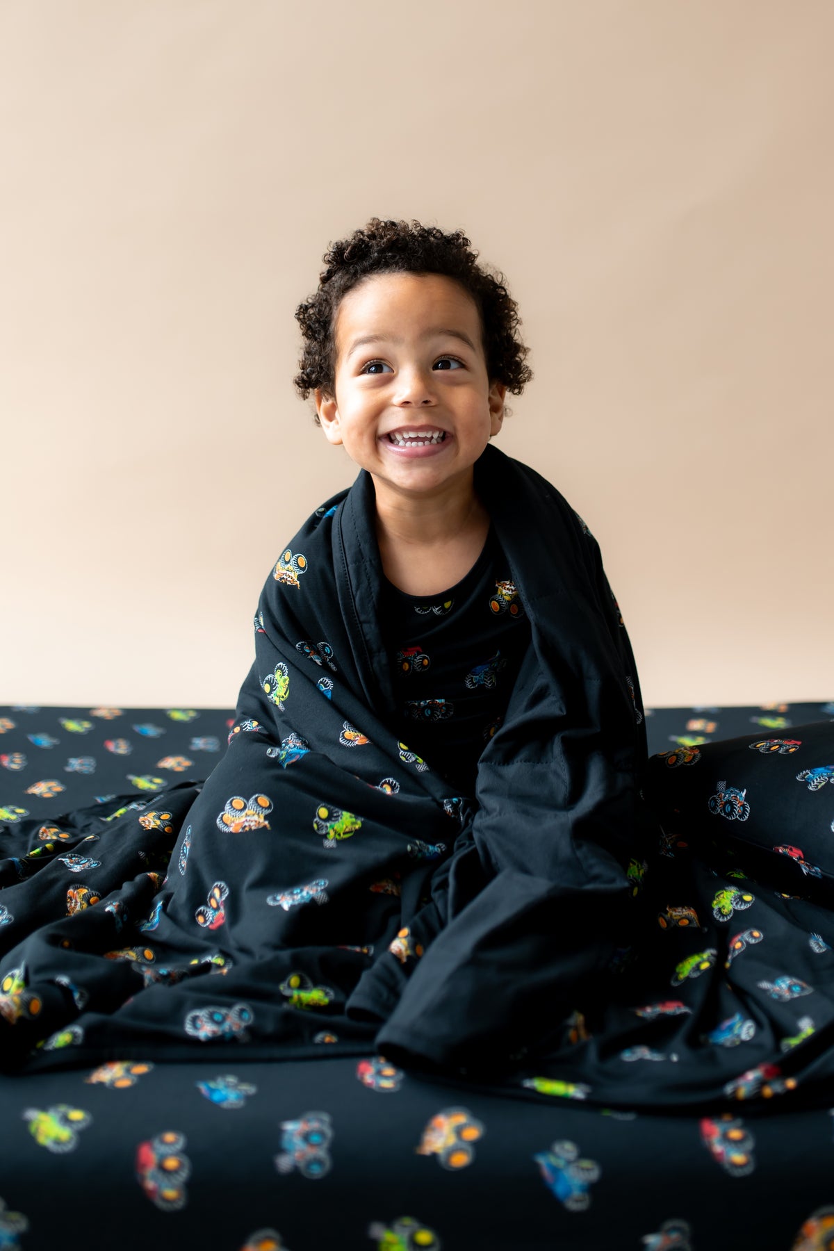 Boy wrapped in black Toddler Blanket in Monster Truck print sitting on similar print sheet.