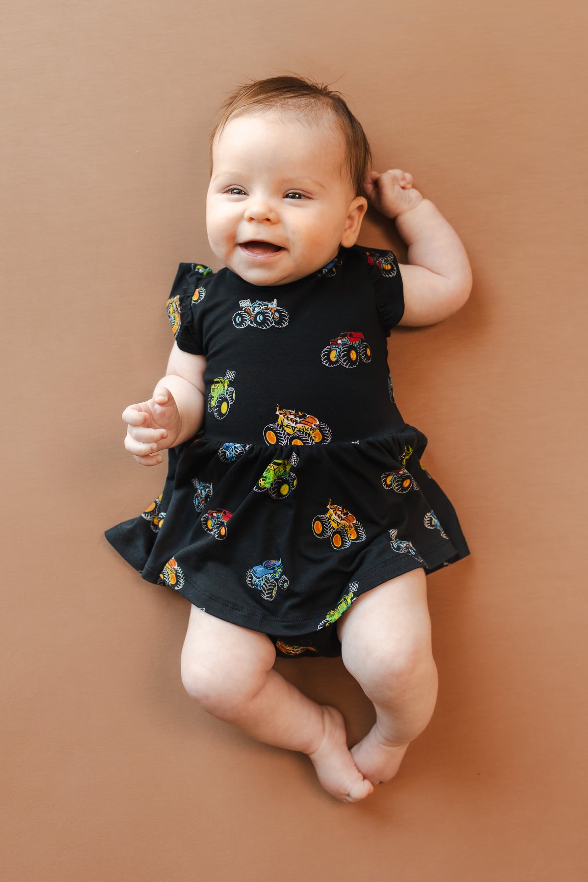 Baby wearing Black Twirl Bodysuit Dress in Monster Truck print on beige background