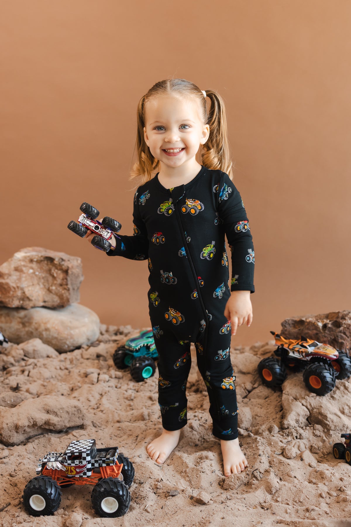Girl holding toy truck wearing black Zippered Romper in Monster Truck print on sand and rocks with toy trucks