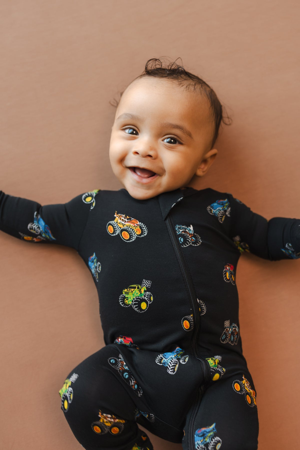 Baby smiling in black Zippered Romper in Monster Truck print on beige background