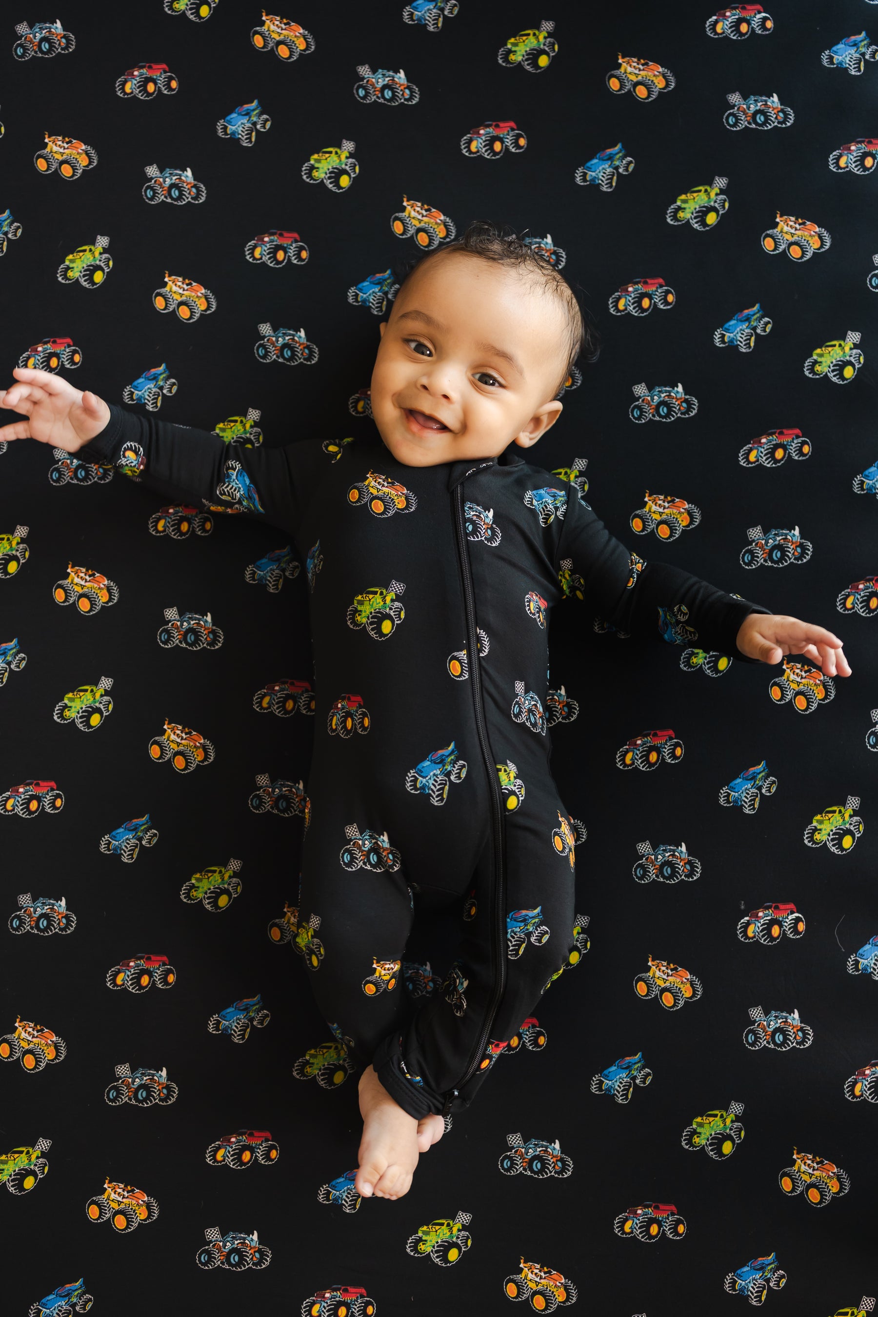 Baby smiling on black crib sheet with Monster truck print wearing similar print Zippered Romper 