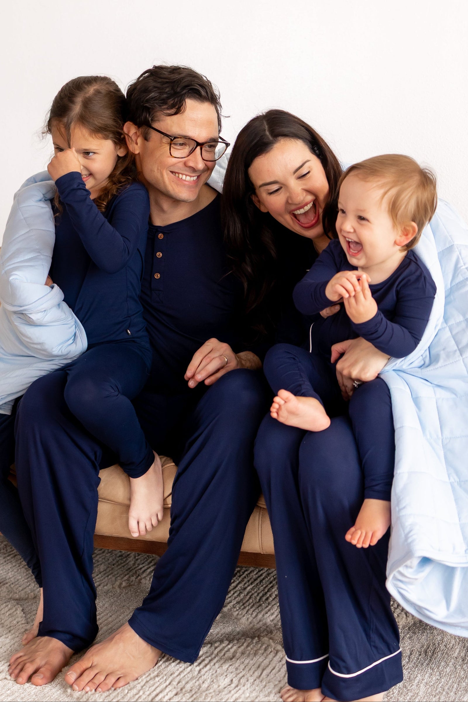 Family of four sitting on a couch wearing matching navy pajamas with a white blanket.