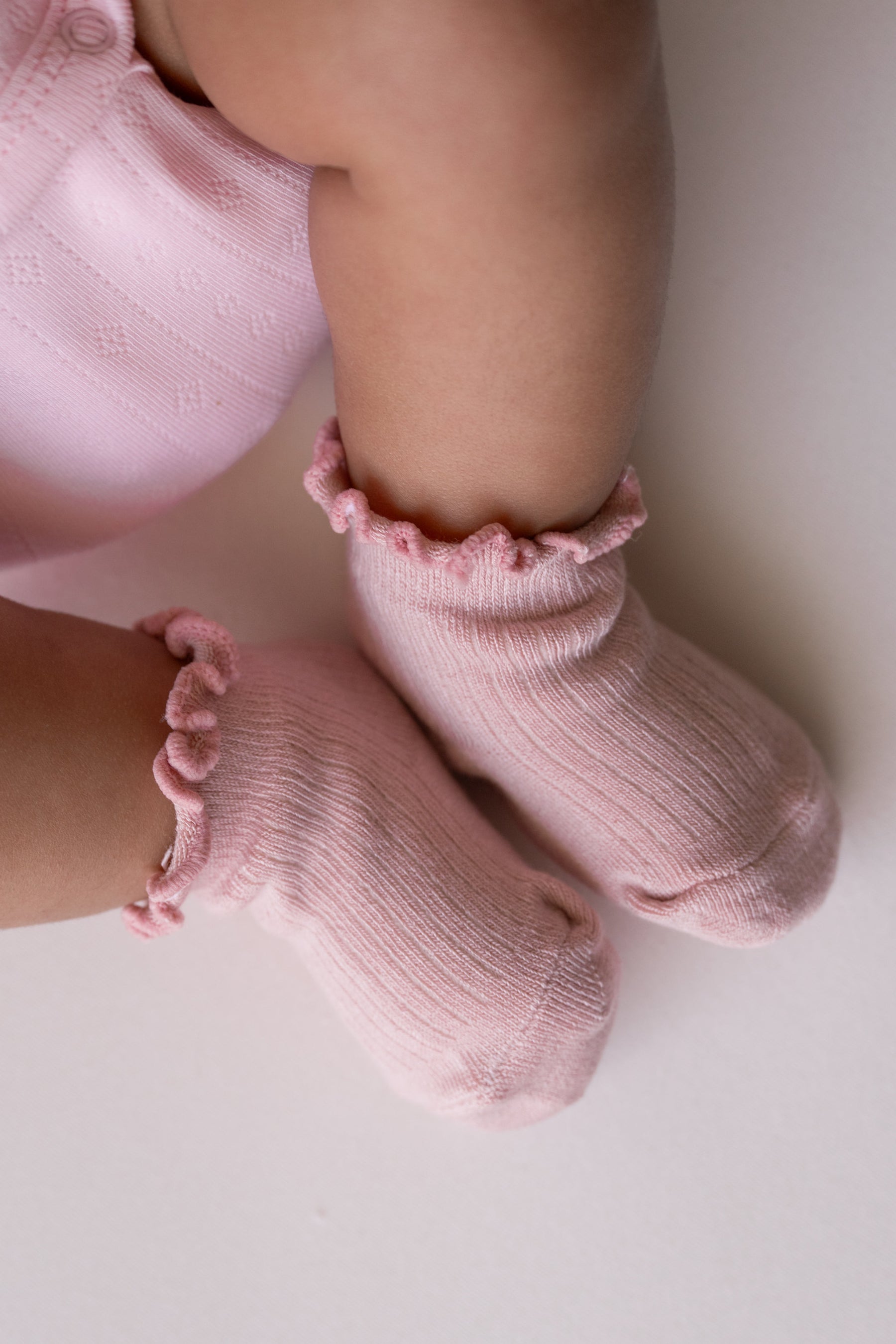 A close-up of a baby's legs wearing light pink ribbed knit socks with ruffled trim, paired with a matching pink outfit on a plain light background.