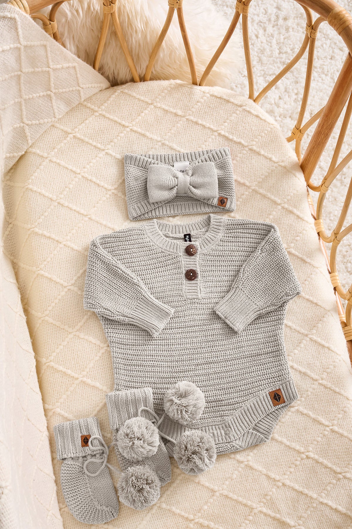 A baby knit outfit set in a wicker crib, featuring a light gray long-sleeve henley bodysuit with three brown buttons, a matching gray bow headband, and gray booties with pom-poms.