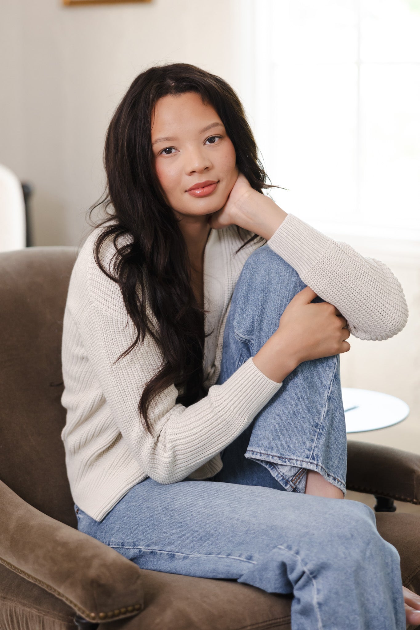 Modal posing in Chunky Knit Women's Cropped Cardigan in Oat over blue jeans while seated on a brown sofa