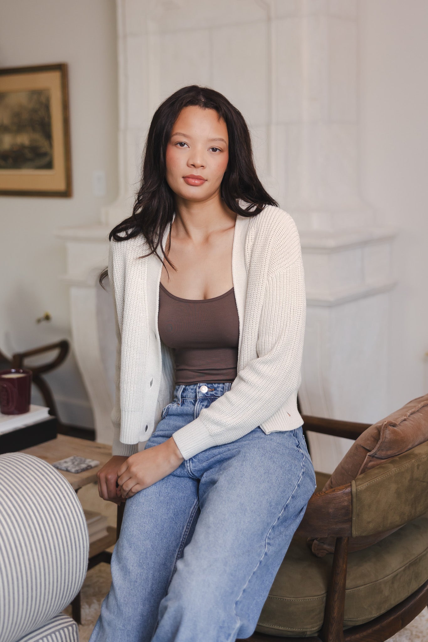 Modal sitting on sofa side wearing Chunky Knit Women's Cropped Cardigan in Oat over espresso camisole and blue jeans