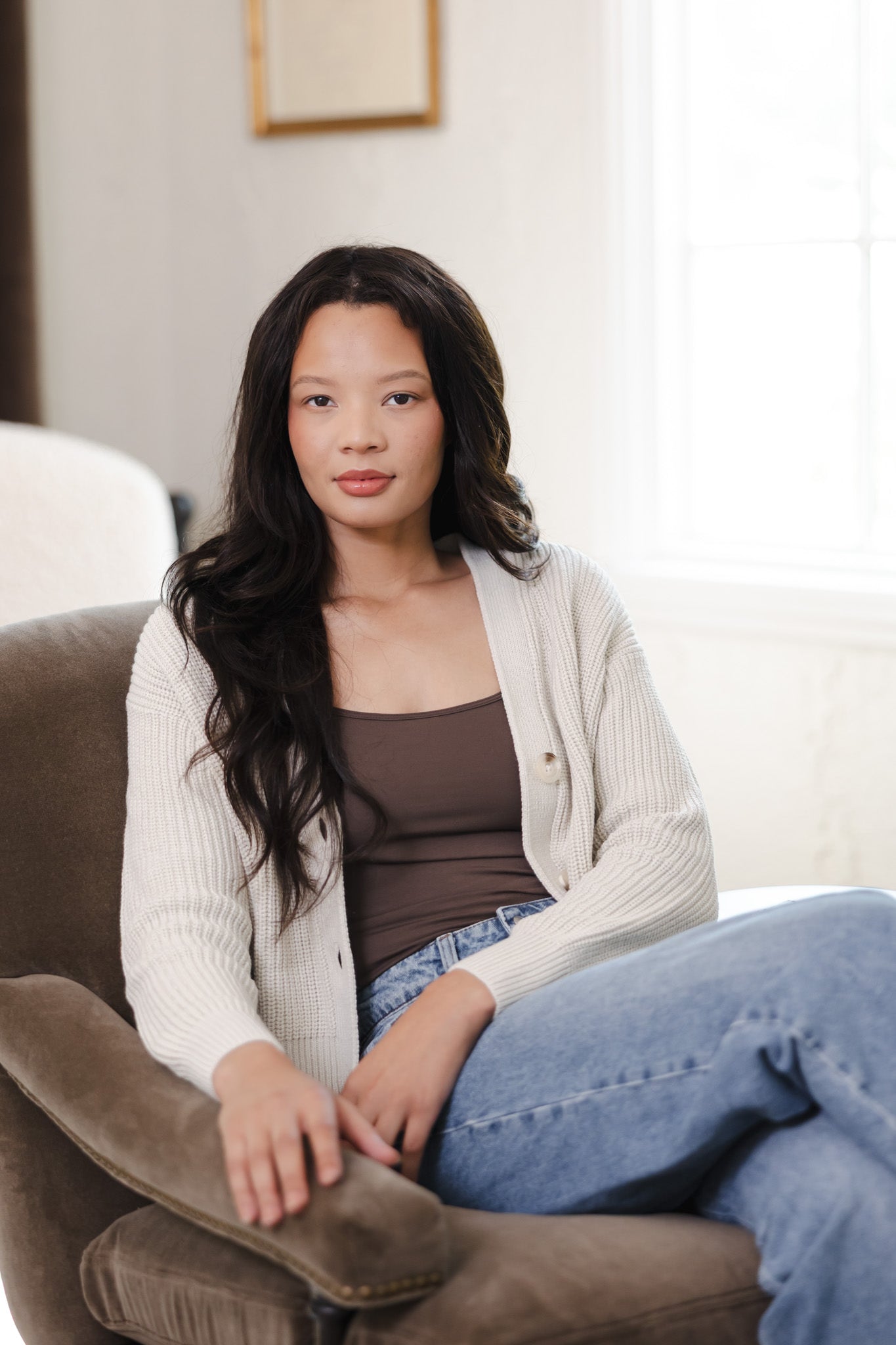 Modal sitting on a brown sofa posing in Chunky Knit Women's Cropped Cardigan in Oat over camisole in espresso and blue jeans