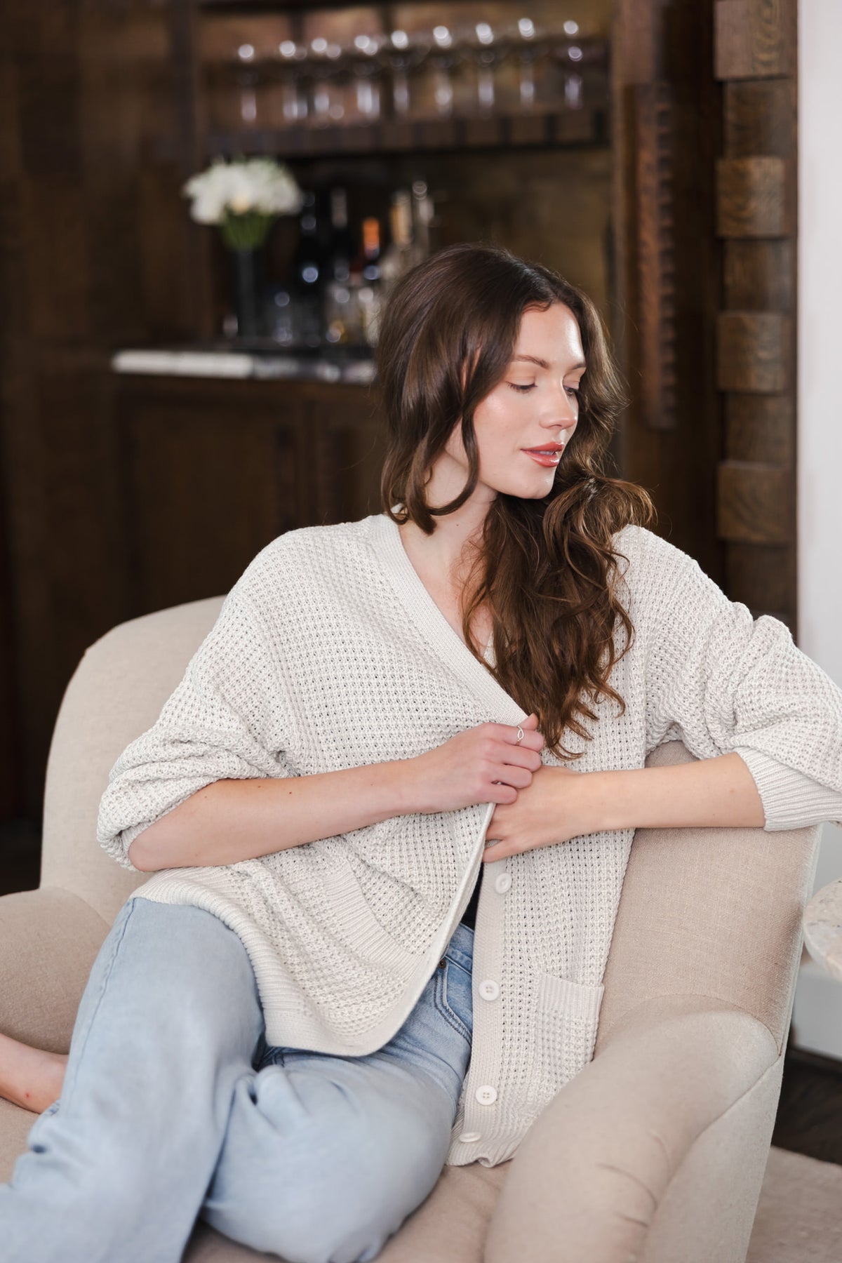 Modal wearing Chunky Knit Women's Waffle Cardigan in Oat over a blue jeans sitting on a sofa