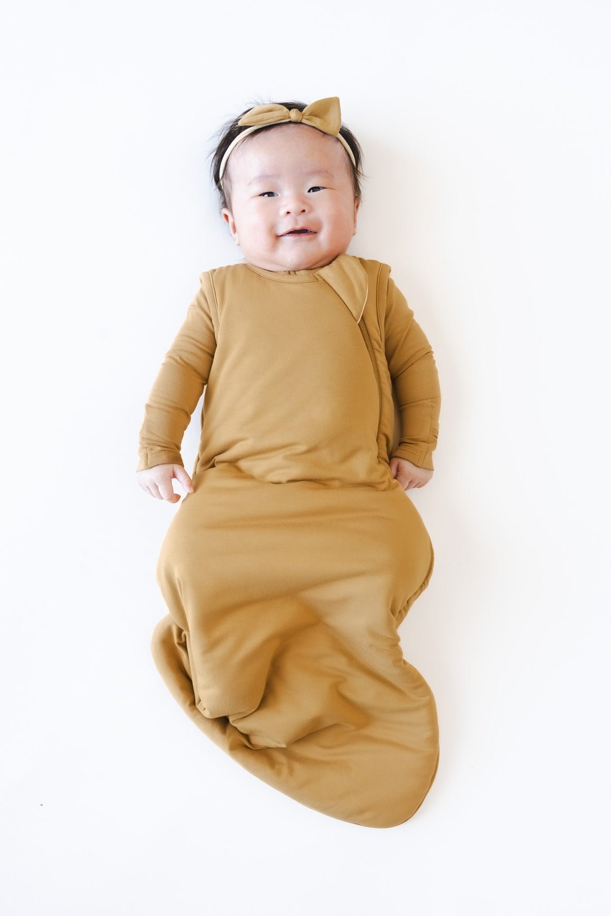 A baby lying on a white background, wearing a mustard yellow sleep sack and bow 