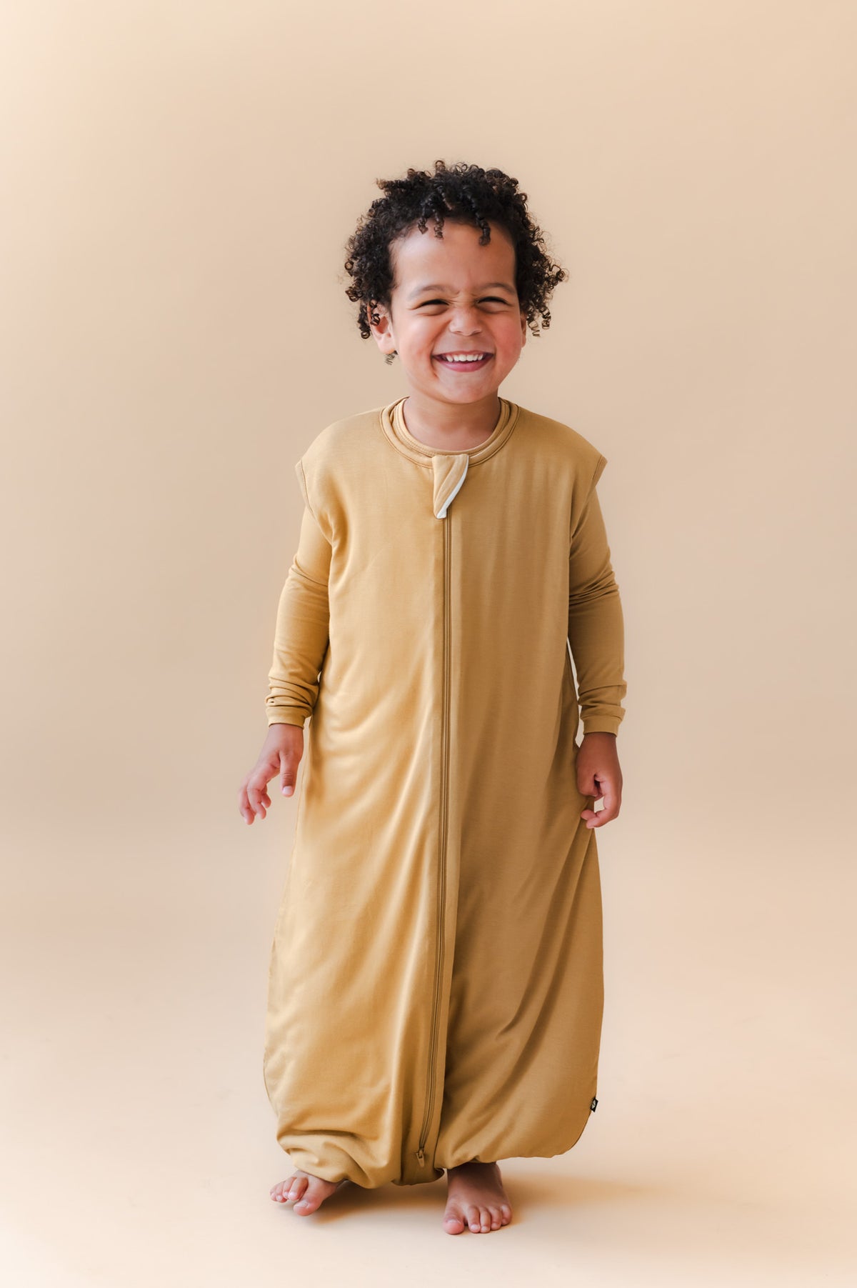 A smiling toddler wearing a mustard yellow full-length zip-up sleep sack walker, standing against a solid beige studio background.