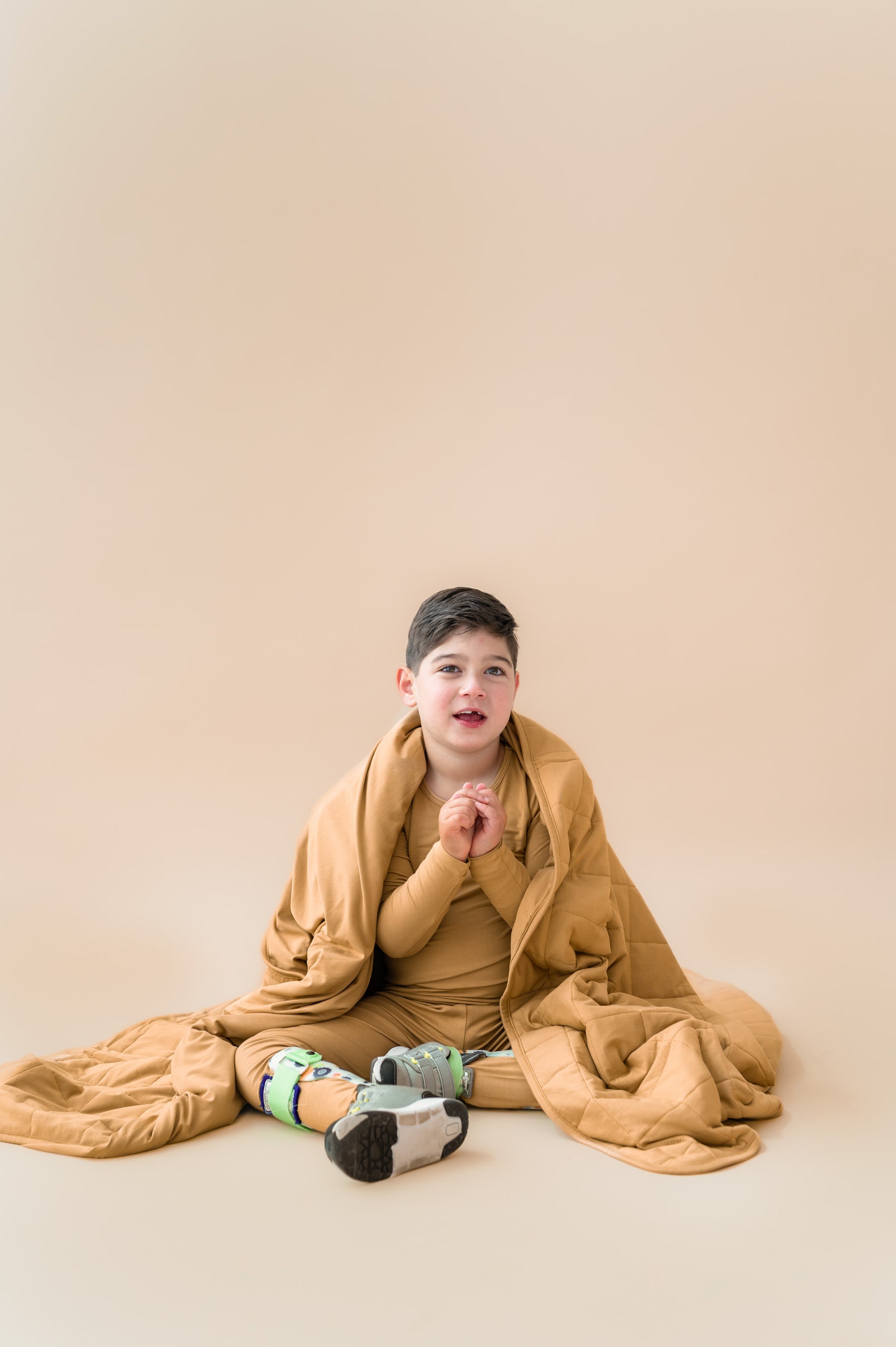 Child covered with Youth Blanket in Ochre 2.5 wearing a similar color long sleeve pajama set on a beige background