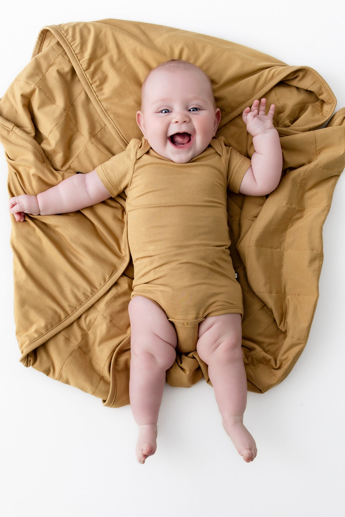 Baby lying over Baby Blanket in Ochre wearing a similar color bodysuit on a white background