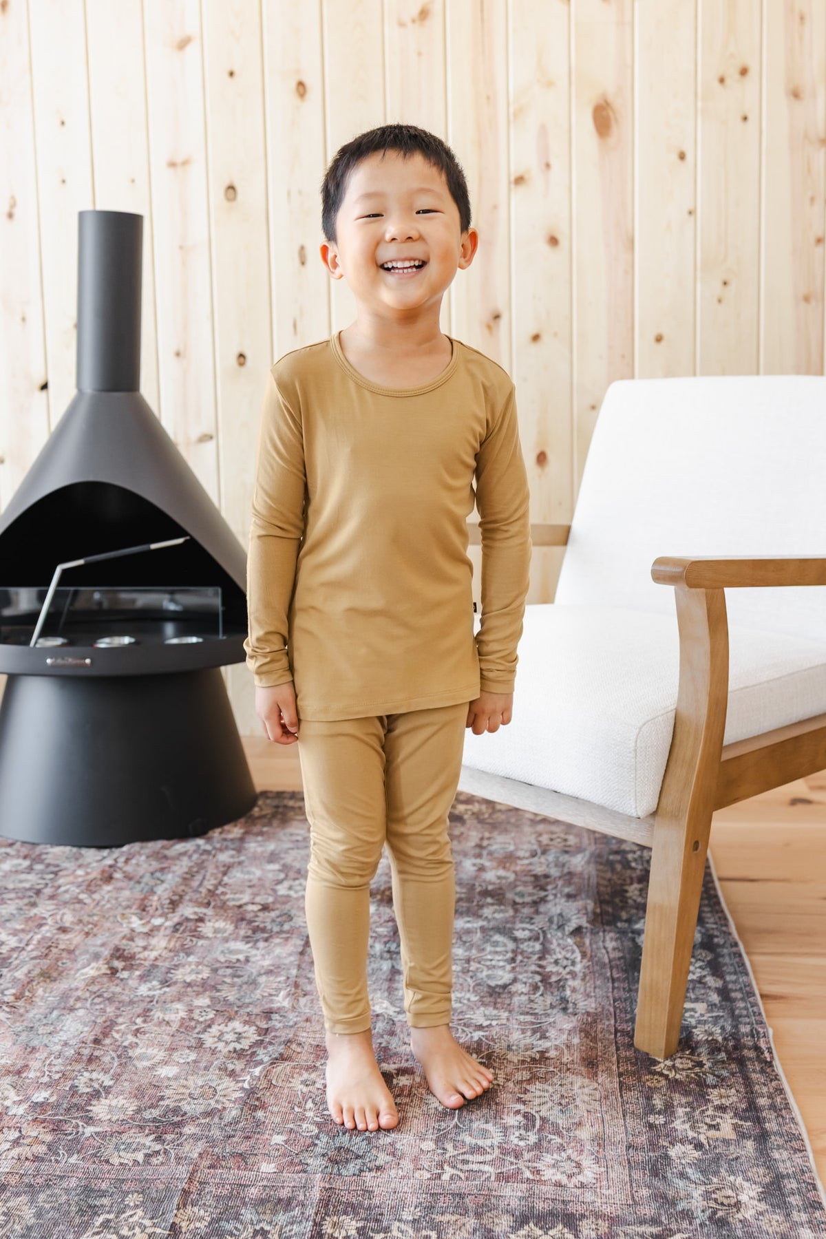 A young boy smiling and standing barefoot indoors, wearing a mustard pajama set. 